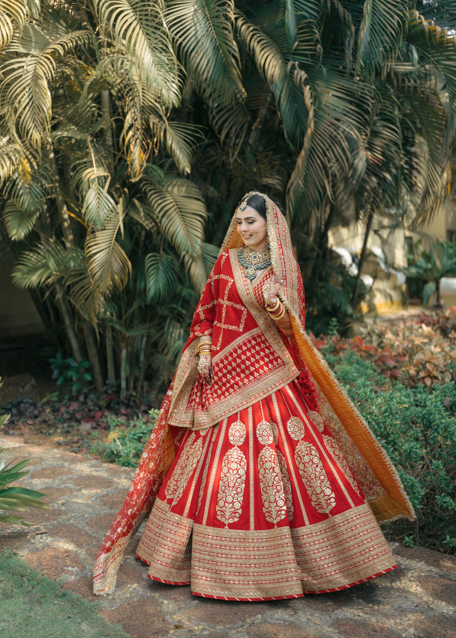 Traditional red bridal lehenga with intricate gold embroidery and embellishments photographed against a palm garden backdrop.