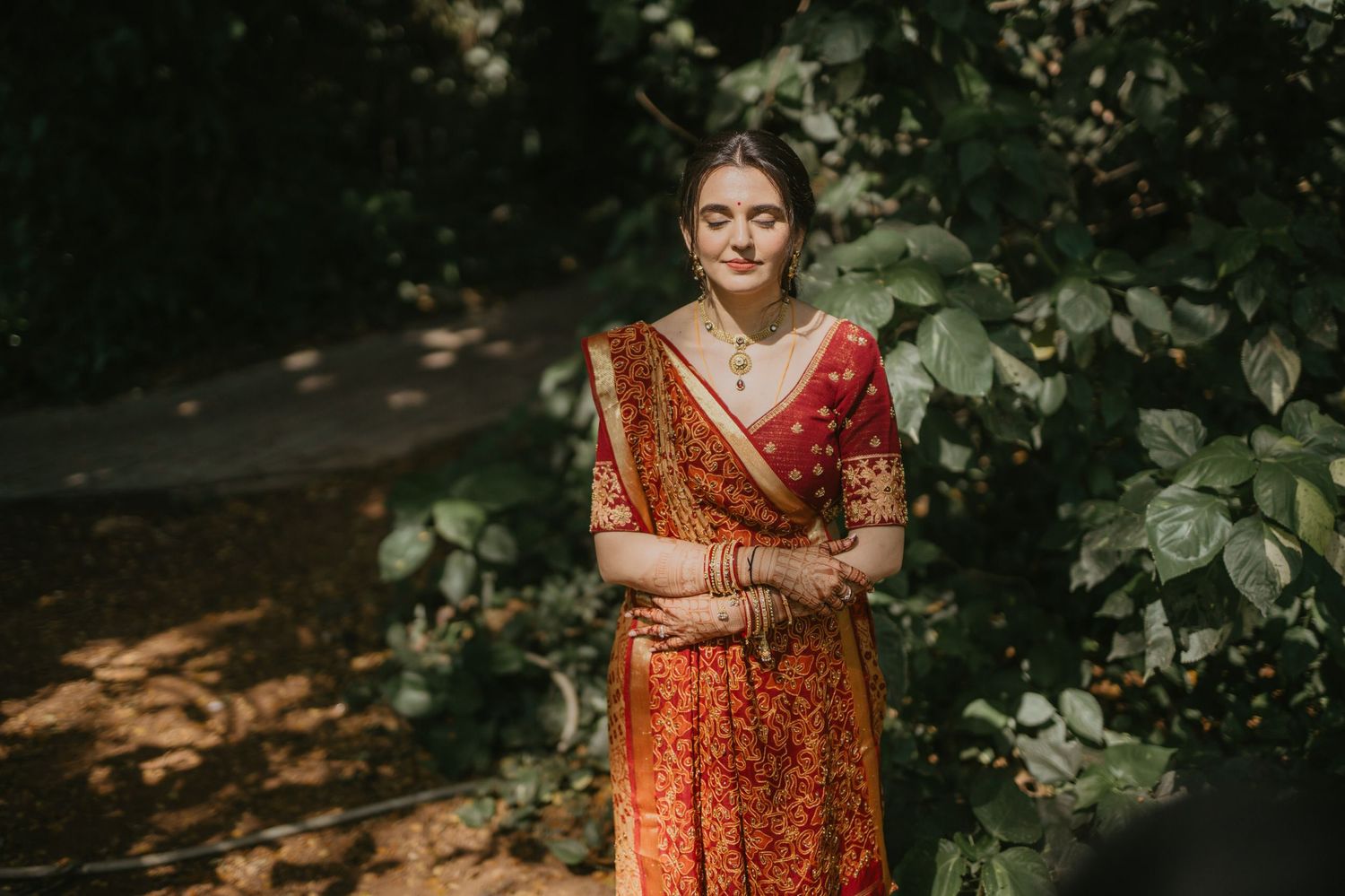 Woman in traditional red and orange Indian sari stands elegantly in dappled sunlight among green foliage.