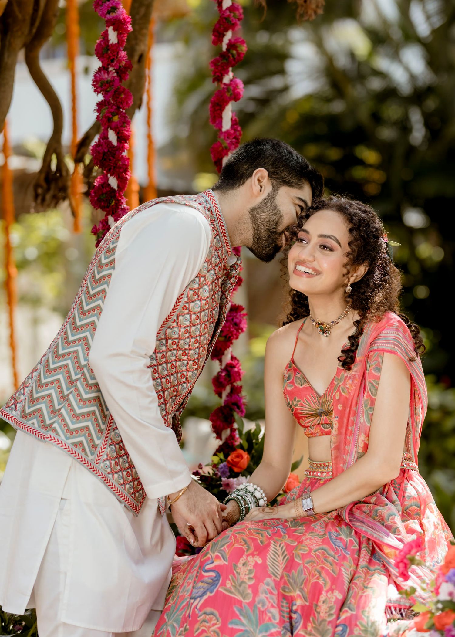 A romantic couple in traditional Indian attire poses under red floral decorations at an outdoor photo shoot.