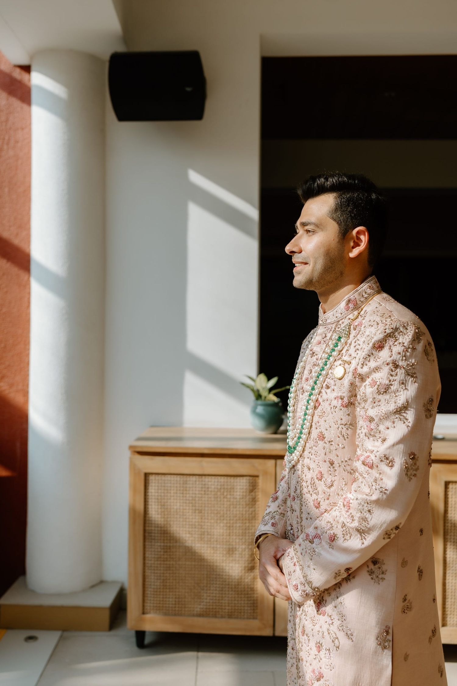 A person in a pink embroidered sherwani stands in front of a rattan chair in a sunlit room.