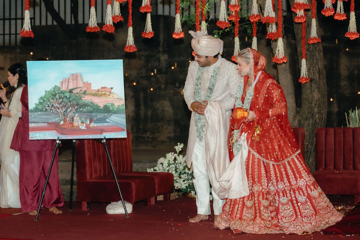 Wedding couple in traditional Indian attire posing under hanging floral decorations with artwork display.