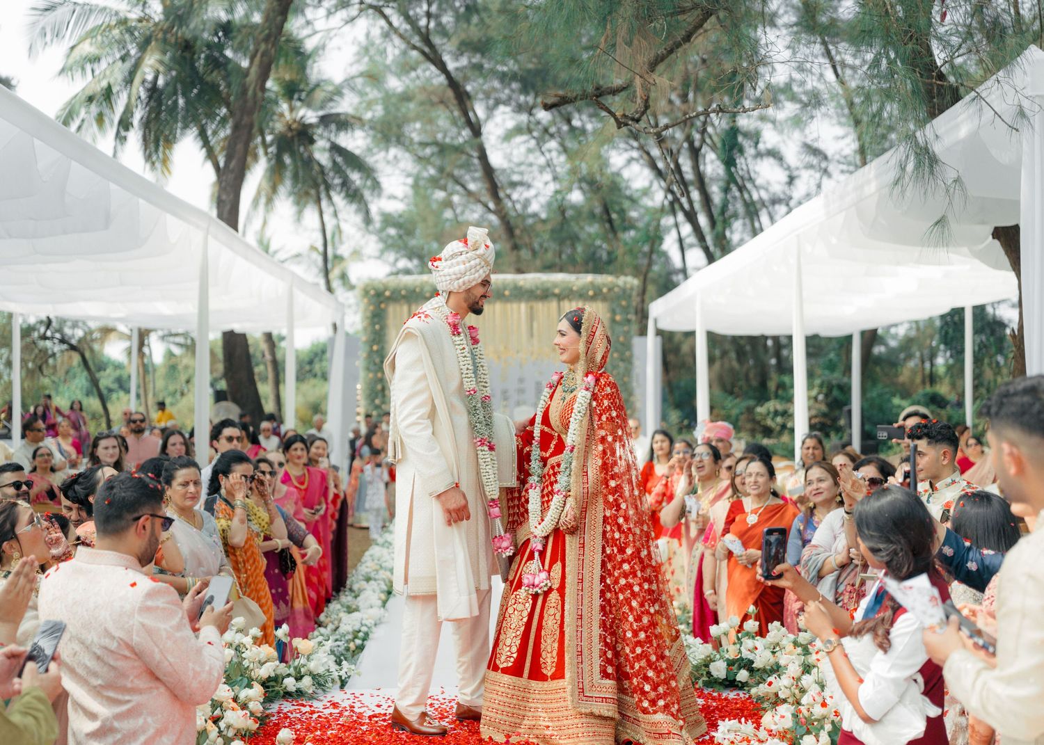 Outdoor wedding ceremony with white canopies and red traditional attire in a palm tree setting.