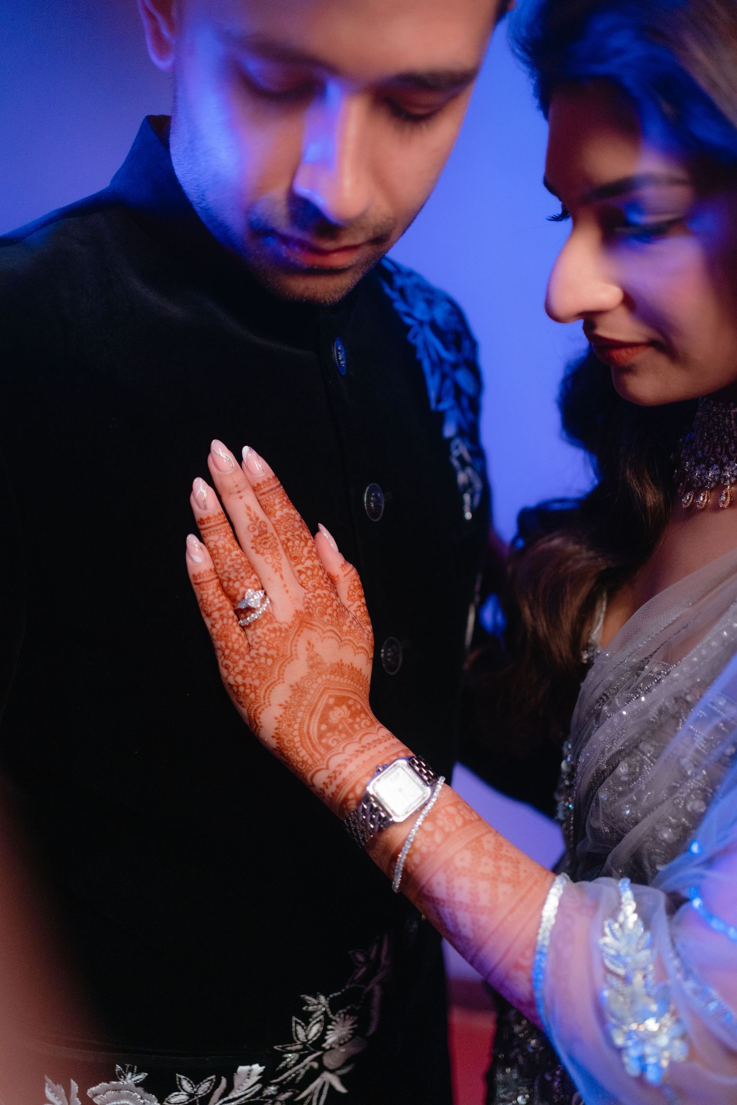 A couple in traditional South Asian attire share an intimate moment with intricate henna designs visible in blue lighting.