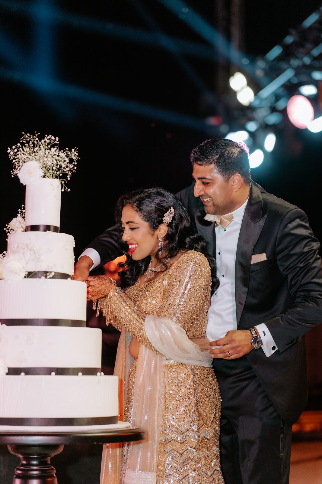 Bride in gold lehenga and groom in black tuxedo cutting a white four-tier wedding cake at an elegant reception.