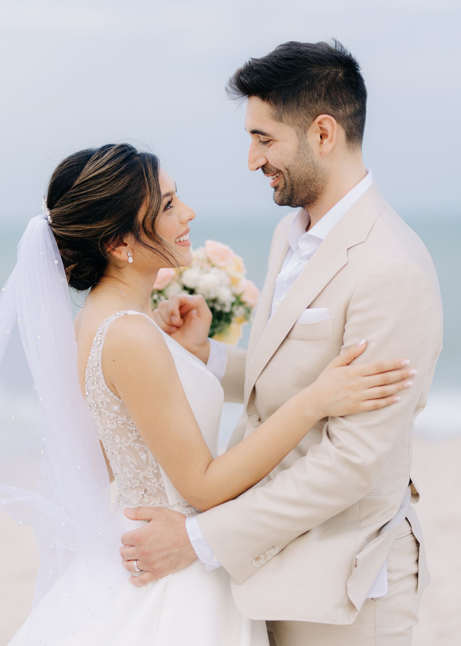 Romantic couple embracing in a tender moment wearing elegant cream and white wedding attire against a soft pastel backdrop
