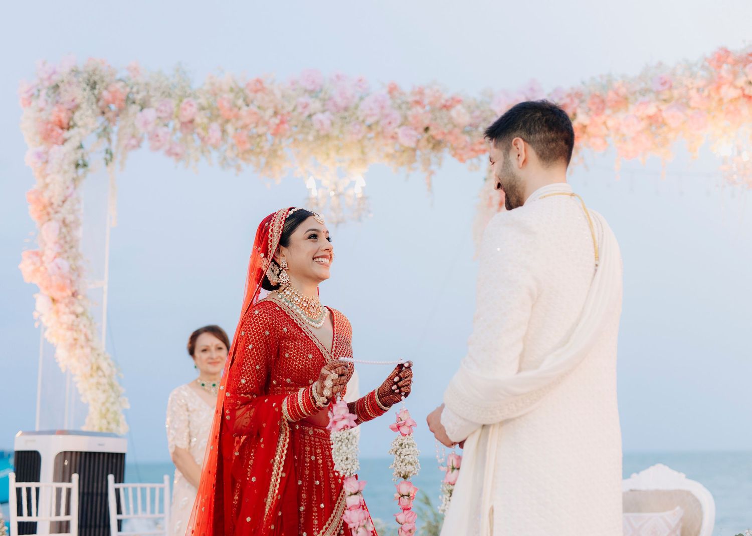 Traditional Indian wedding ceremony takes place under a beautifully decorated floral canopy with ocean in background.