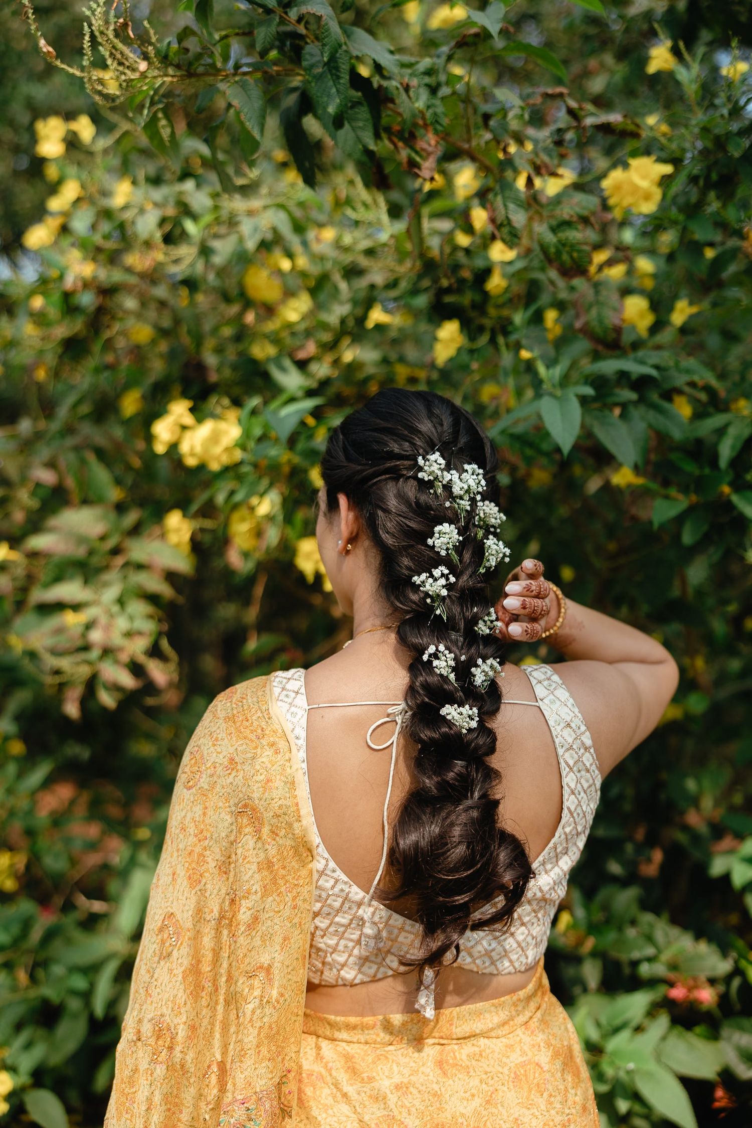 Woman in yellow lehenga with floral braid hairstyle, adorned with white flowers, standing before yellow blooms.