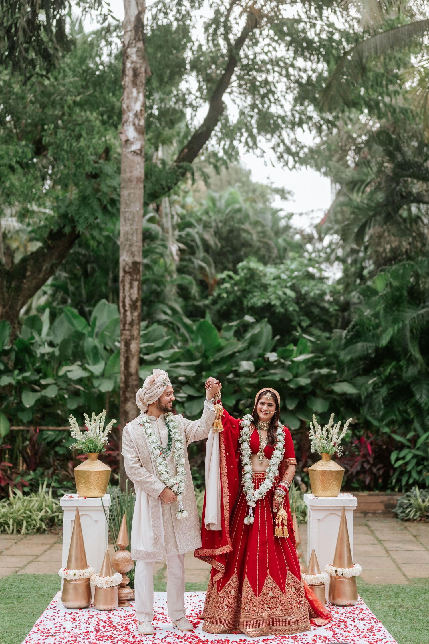 Indian bride in red lehenga and groom in white sherwani hold hands during outdoor Hindu wedding ceremony.