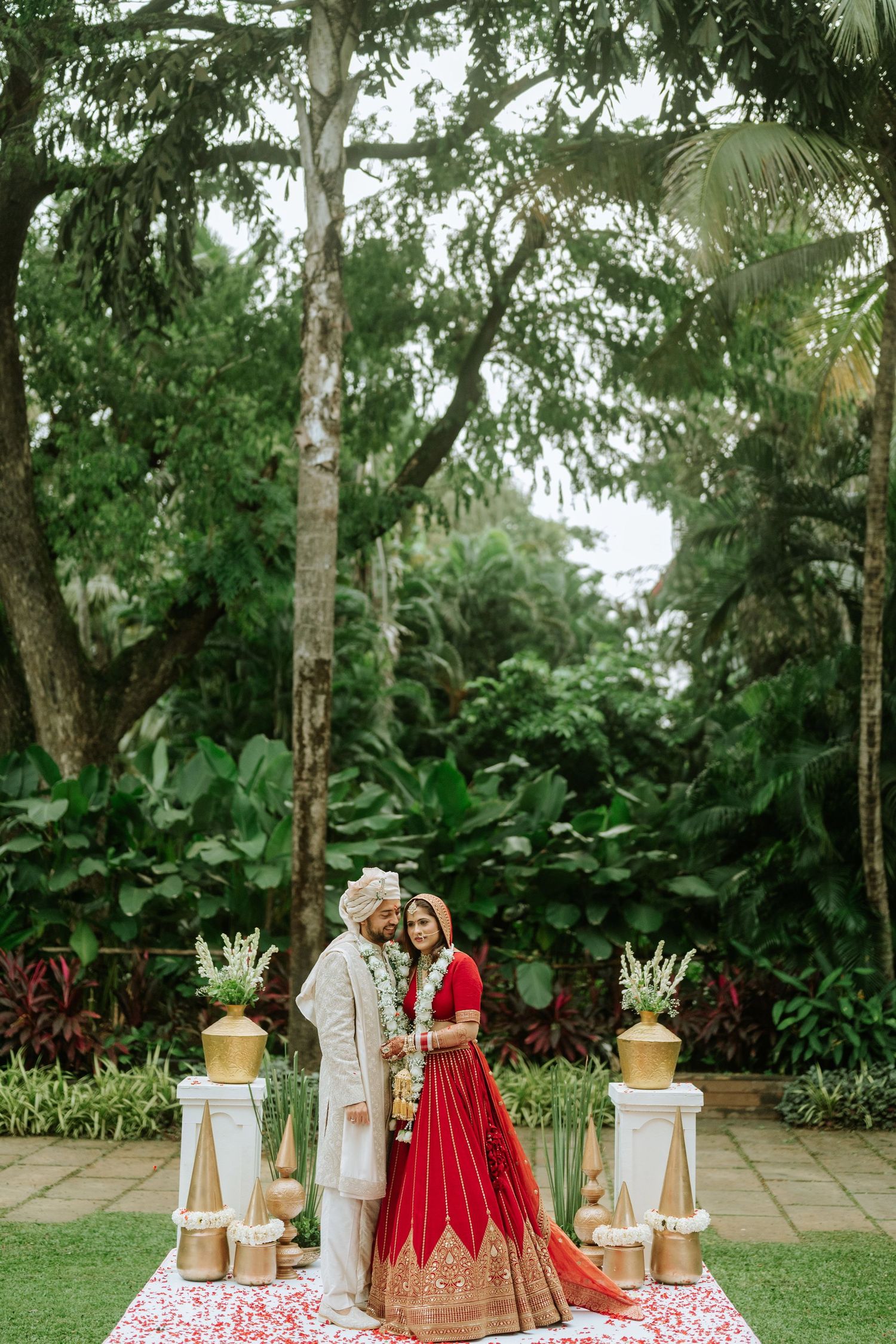 Indian bride in red lehenga and groom in white sherwani pose together at outdoor garden wedding ceremony.