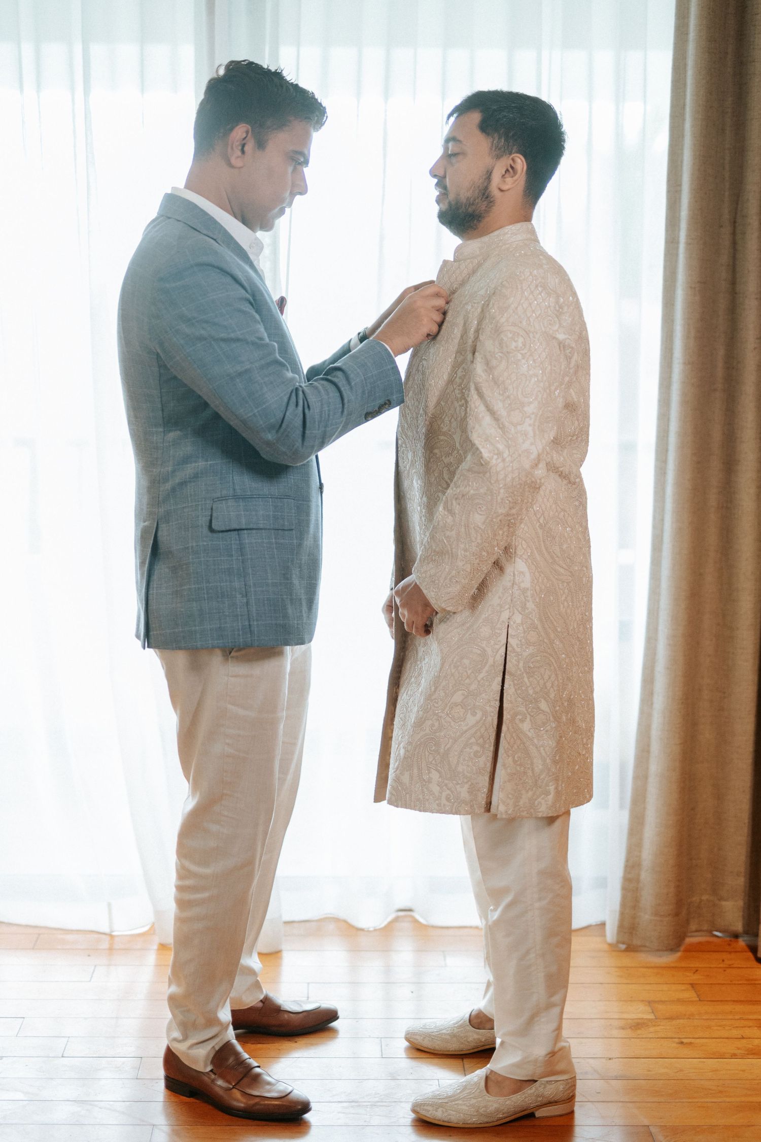 Two men in formal attire help each other prepare for a wedding ceremony indoors.