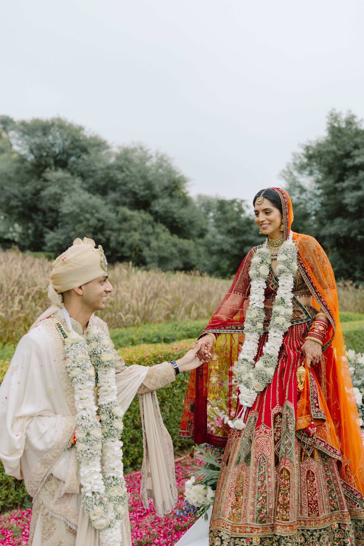 Couple in traditional Indian wedding attire posing in garden with floral garlands.