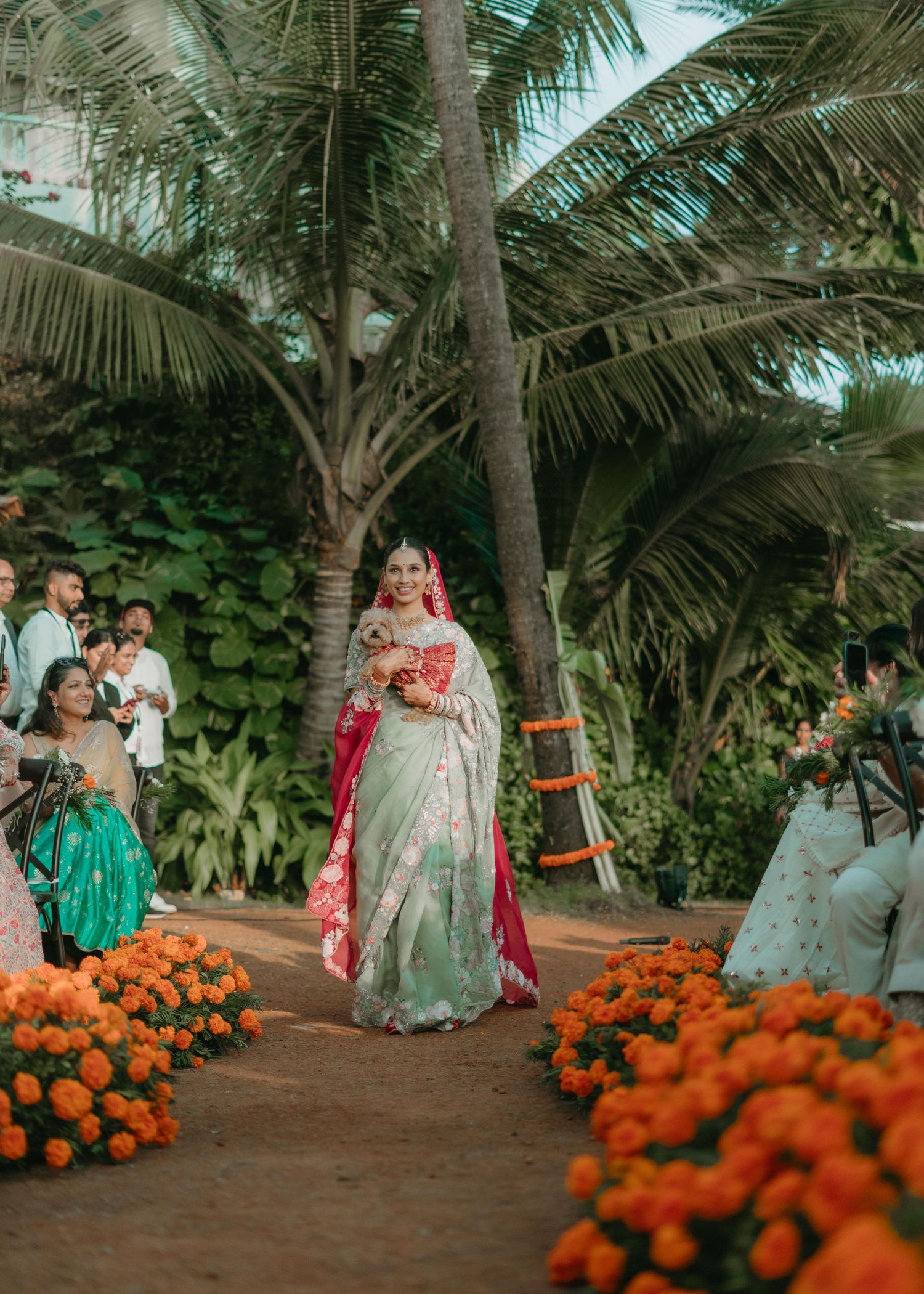 Bride in a white and red saree walks down a floral aisle lined with marigolds at an outdoor Indian wedding.