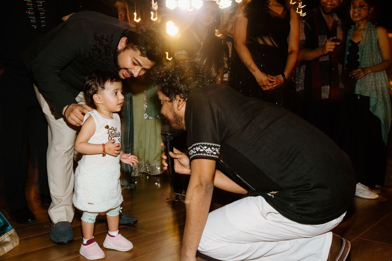 Two men interact warmly with a toddler in pink shoes at an indoor celebration event.