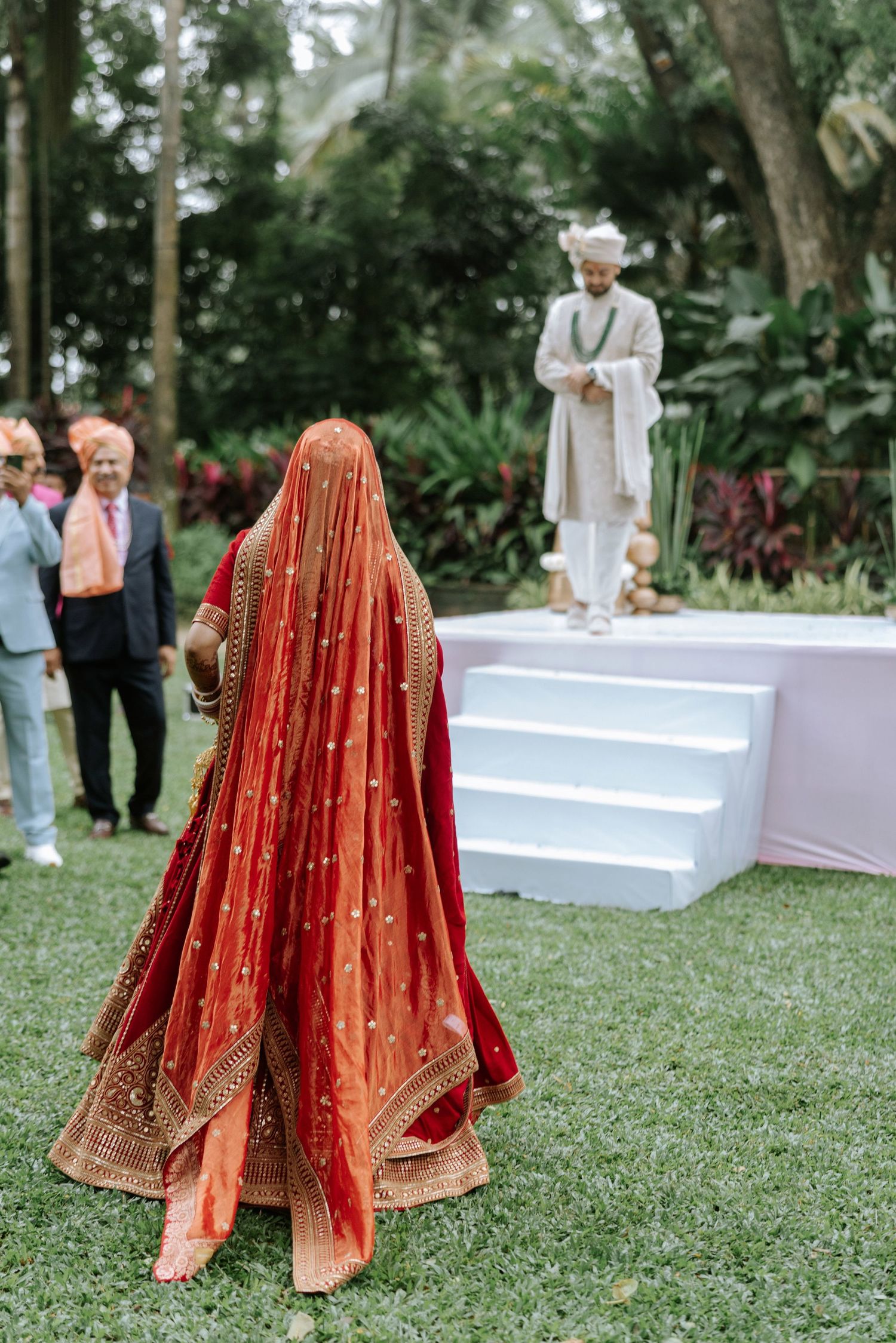 Indian bride in red lehenga walks toward groom in white sherwani at outdoor Hindu wedding ceremony.