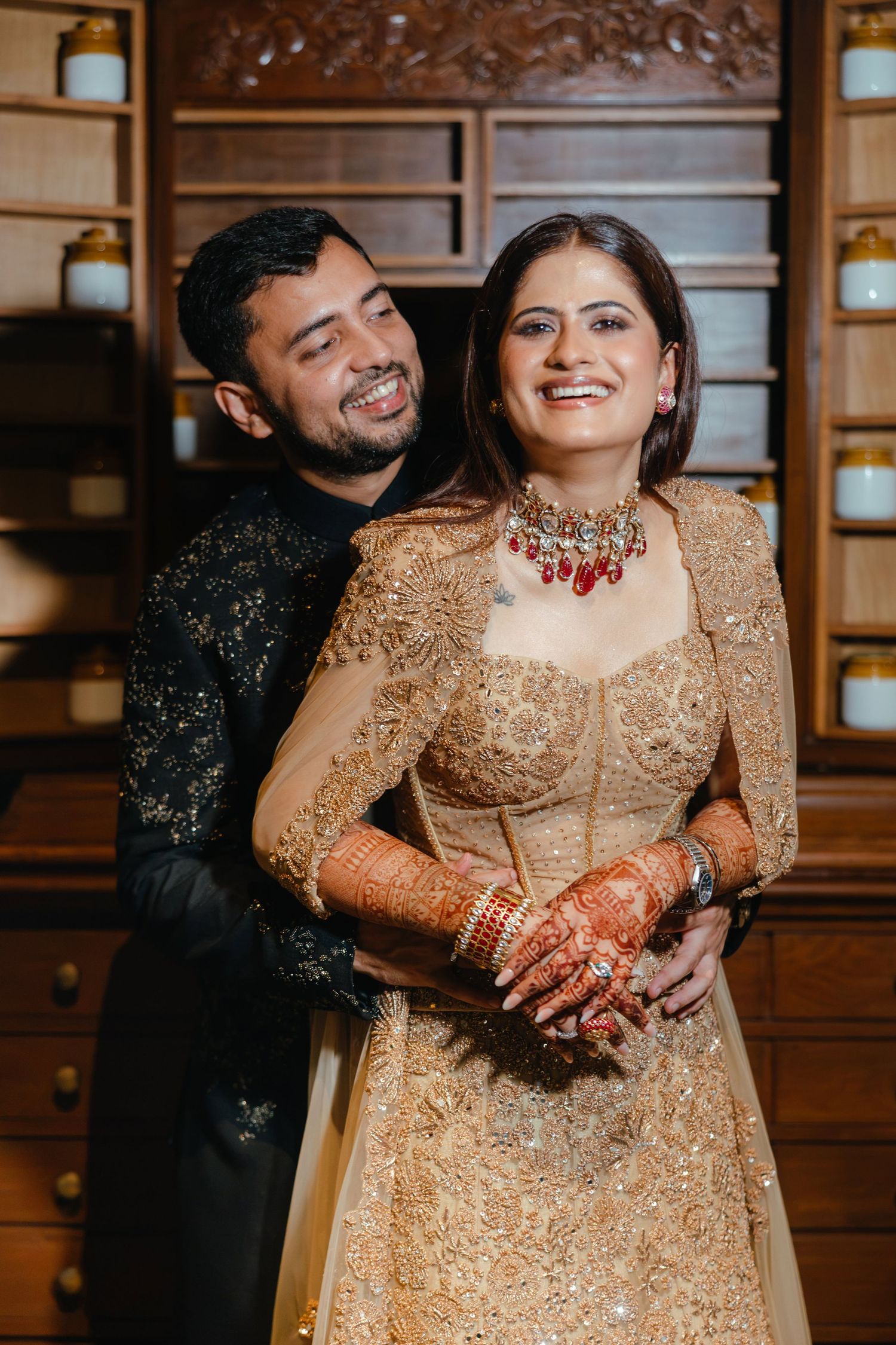 Indian couple in traditional wedding attire, bride in gold lehenga with red jewelry, groom in black sherwani, smiling together.