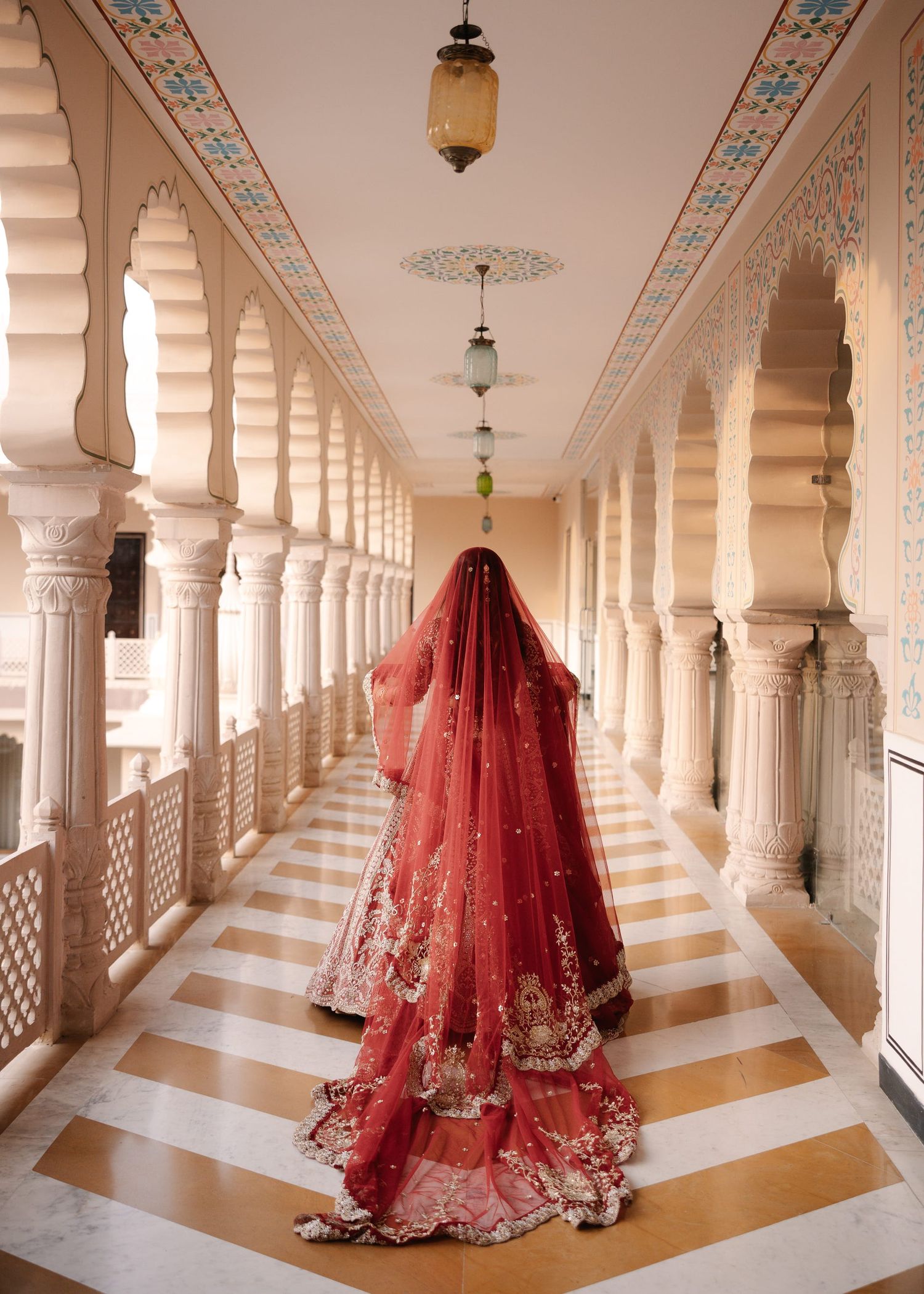 Indian bride in red lehenga with embroidered dupatta walks through ornate marble palace corridor.