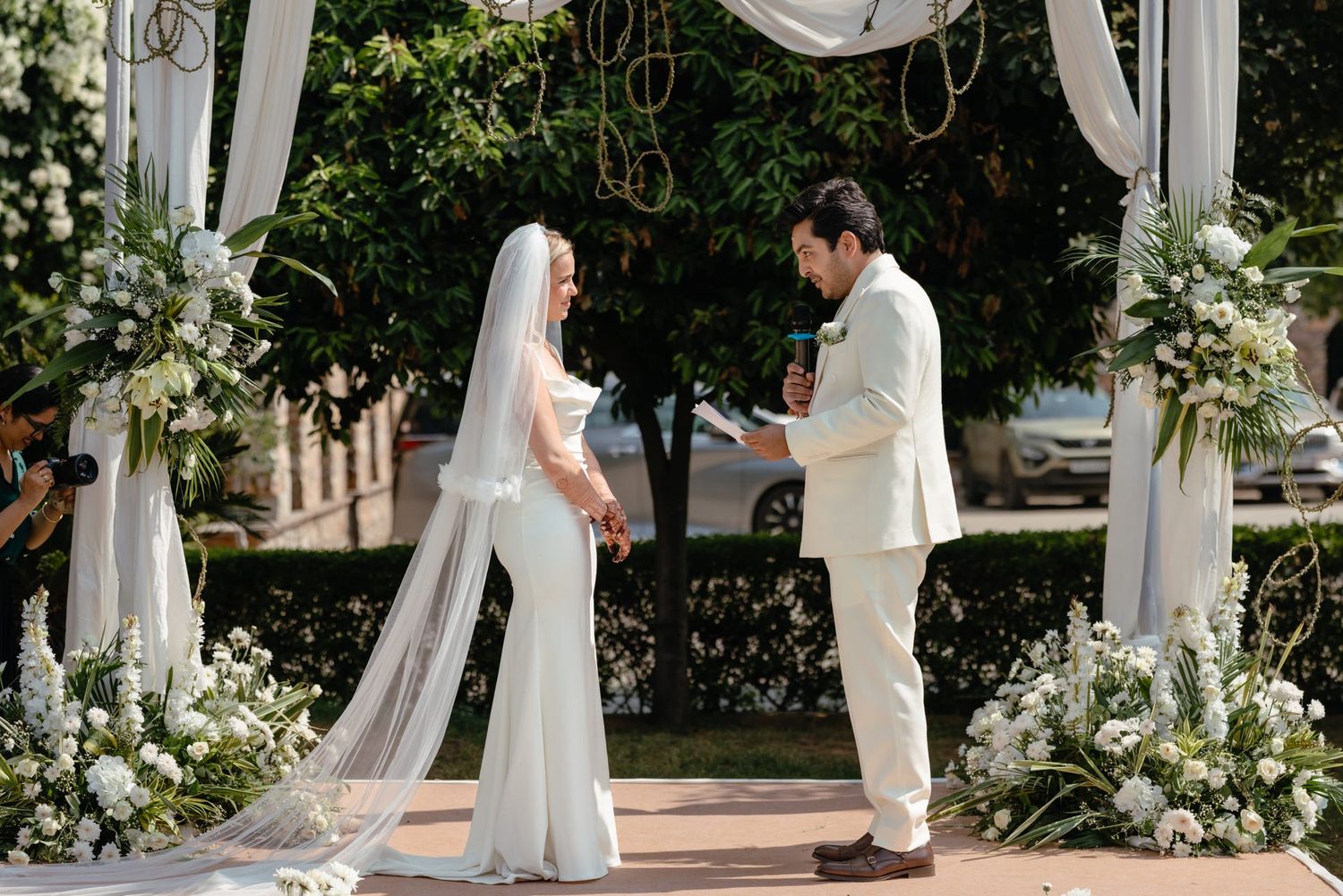 Wedding ceremony setup with white draped pillars and floral arrangements in garden setting.
