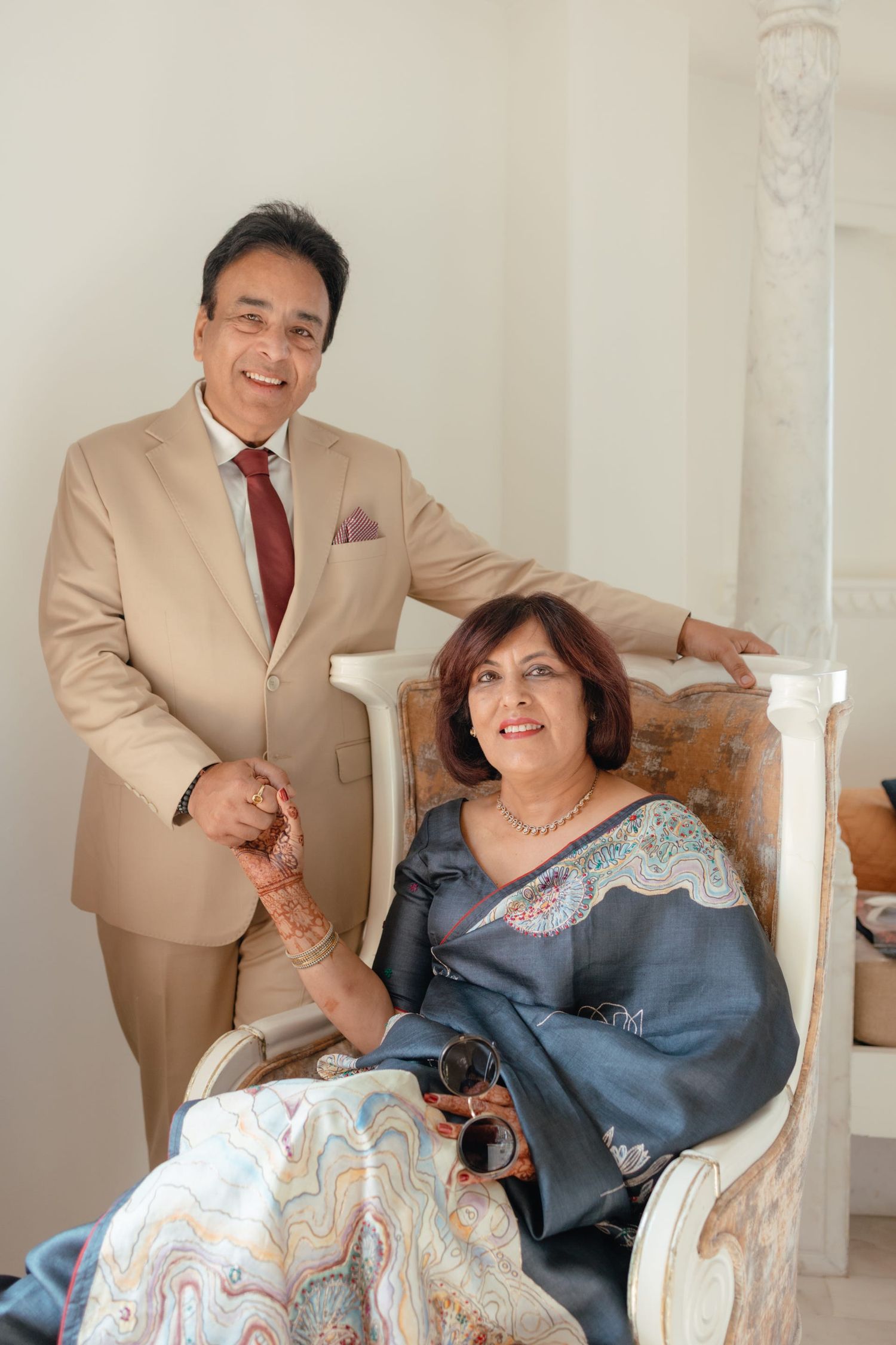 Elegant couple in formal attire share a tender moment in an upholstered chair against a light wall.