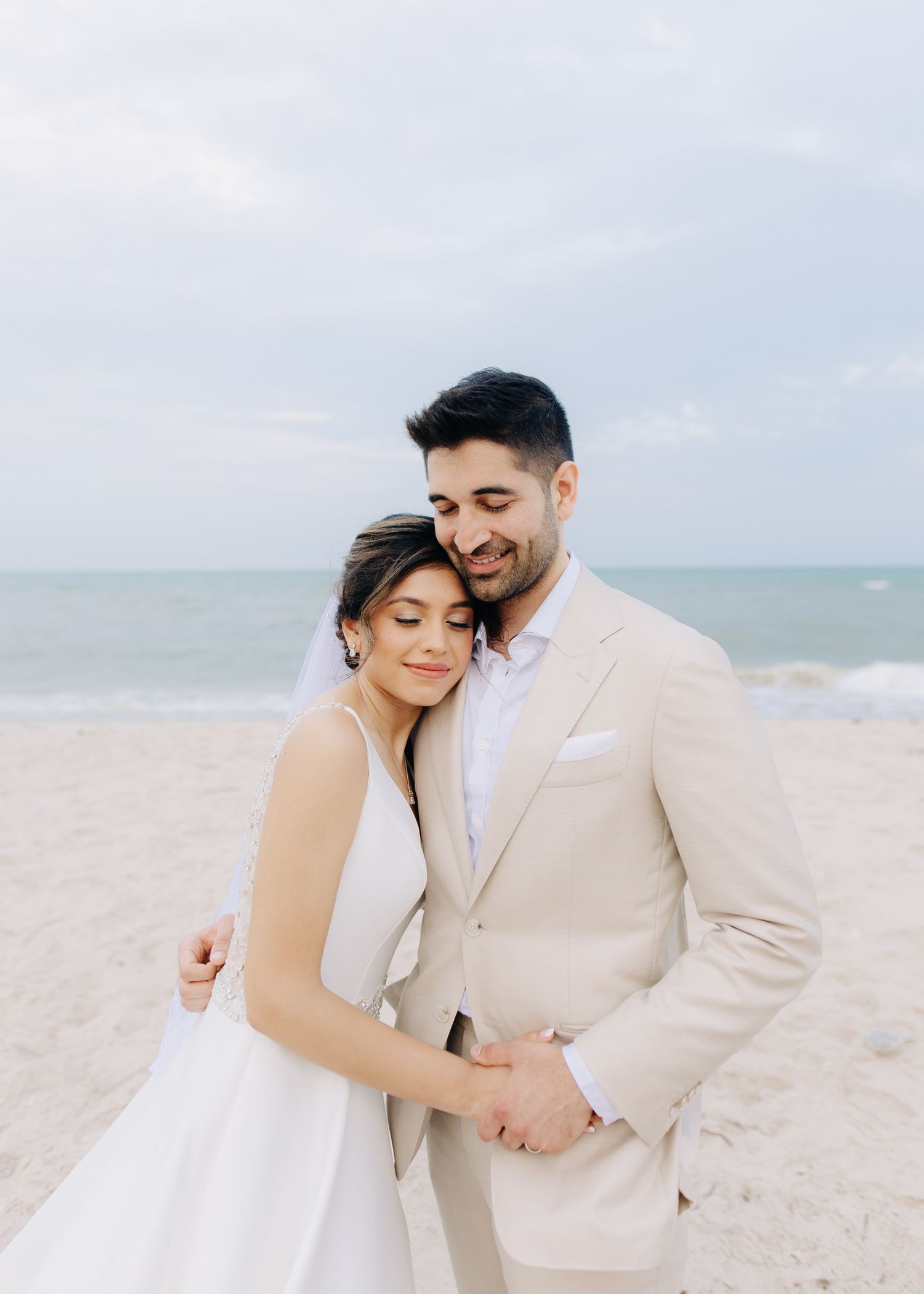 A romantic couple in wedding attire embrace on a serene beach with ocean waves in the background.