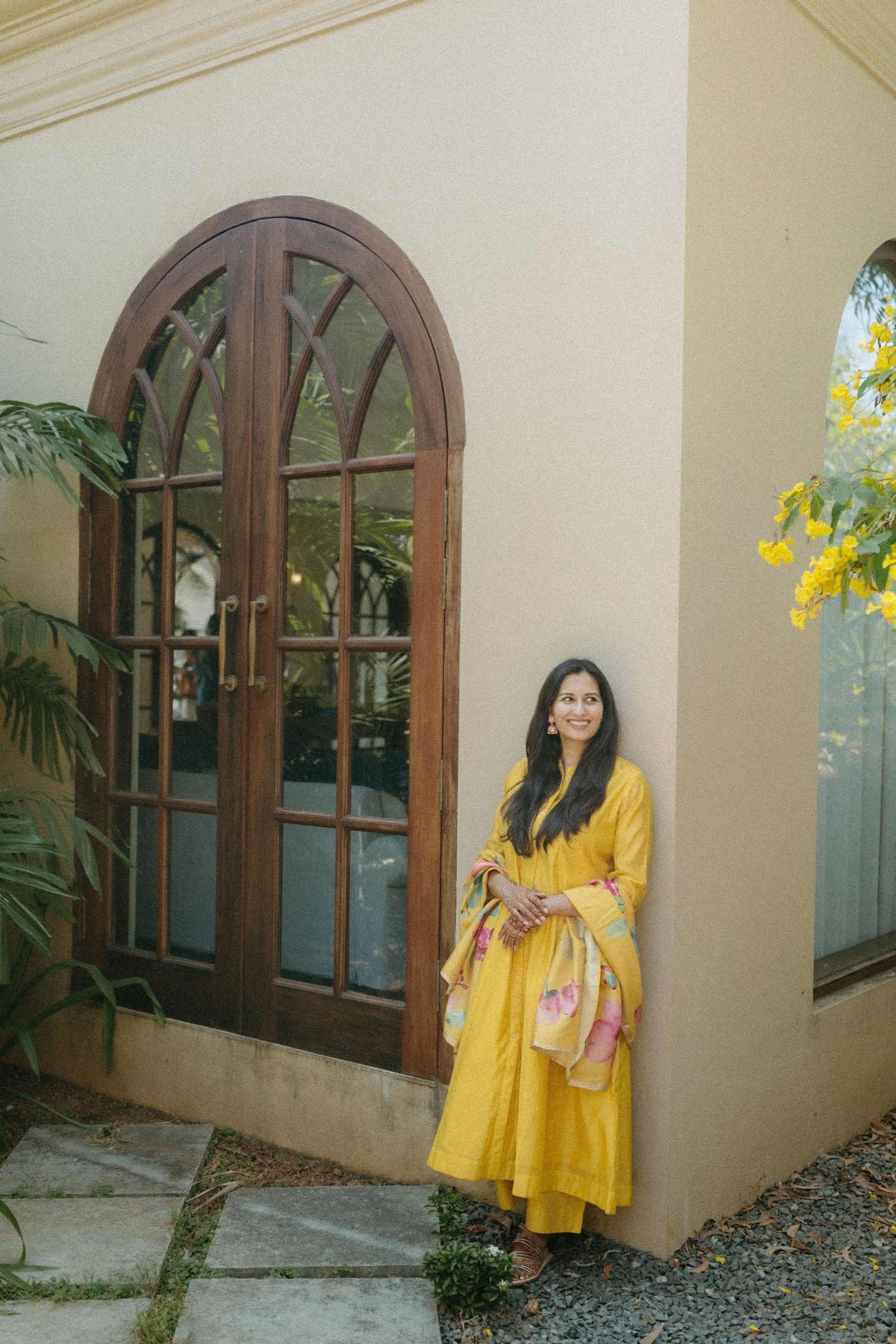 Person in vibrant yellow dress stands by arched wooden door with glass panels at corner of beige building.