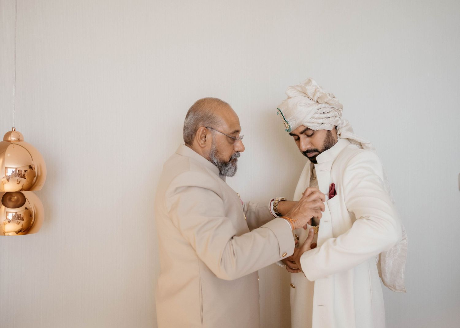 Two people in white and beige outfits share a tender moment while adjusting buttons on a formal outfit.