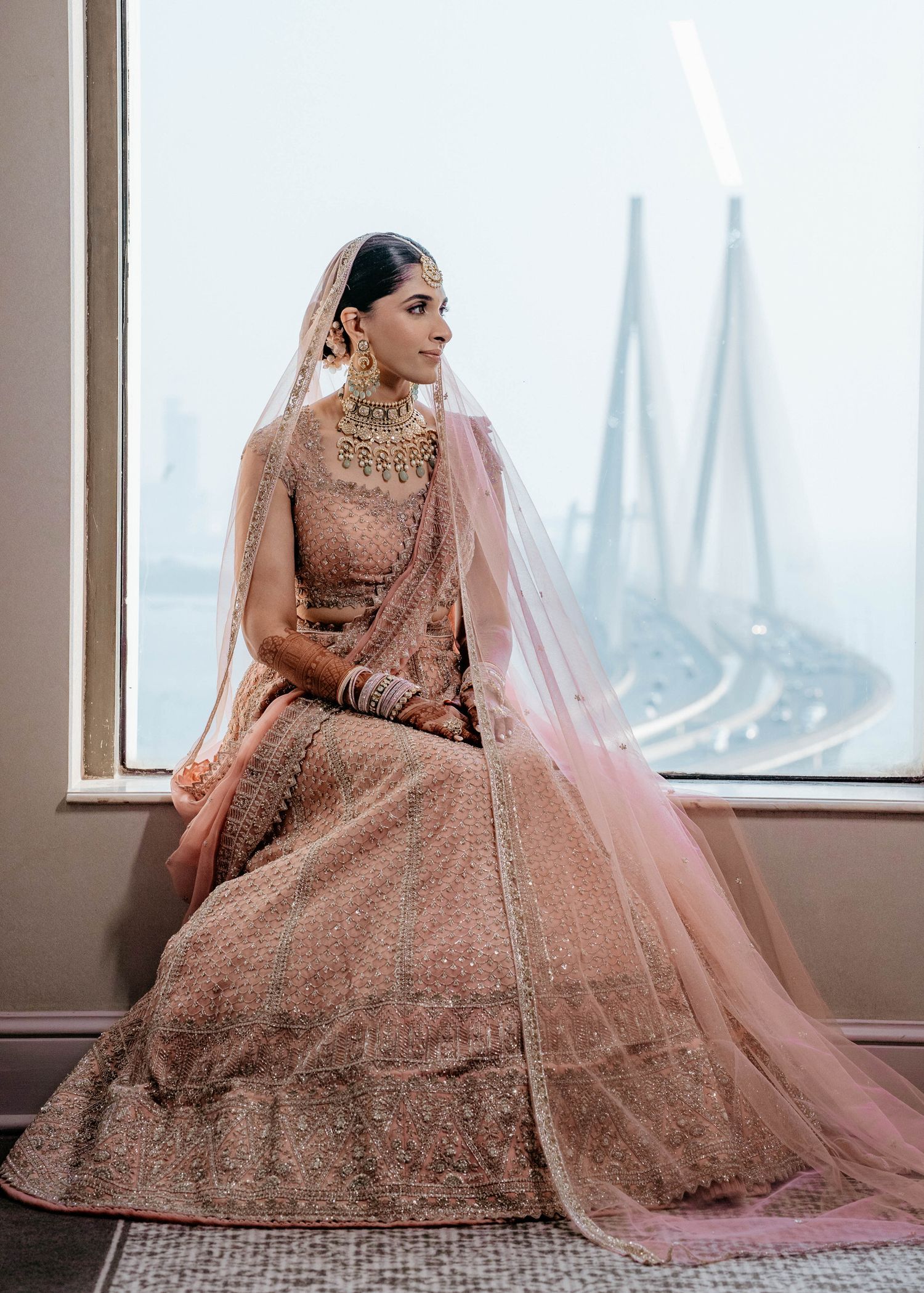 A bride in an elegant rose gold lehenga choli with intricate embroidery poses near floor-to-ceiling windows with city views.