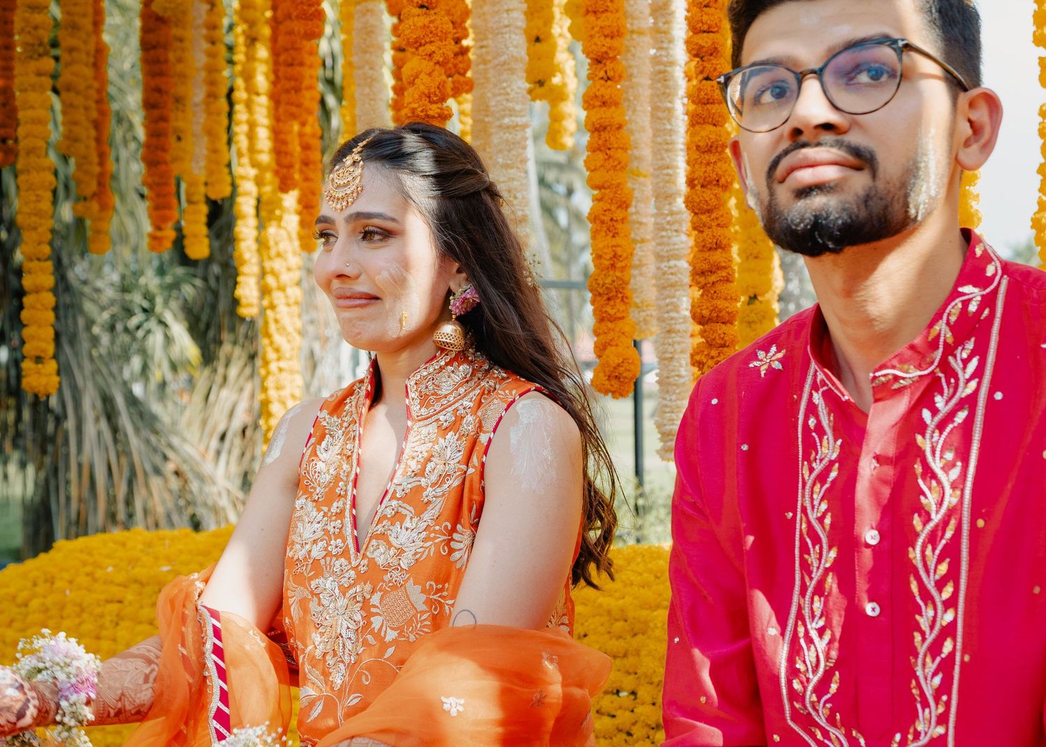Two people in traditional Indian wedding attire sit together surrounded by bright marigold flower decorations.