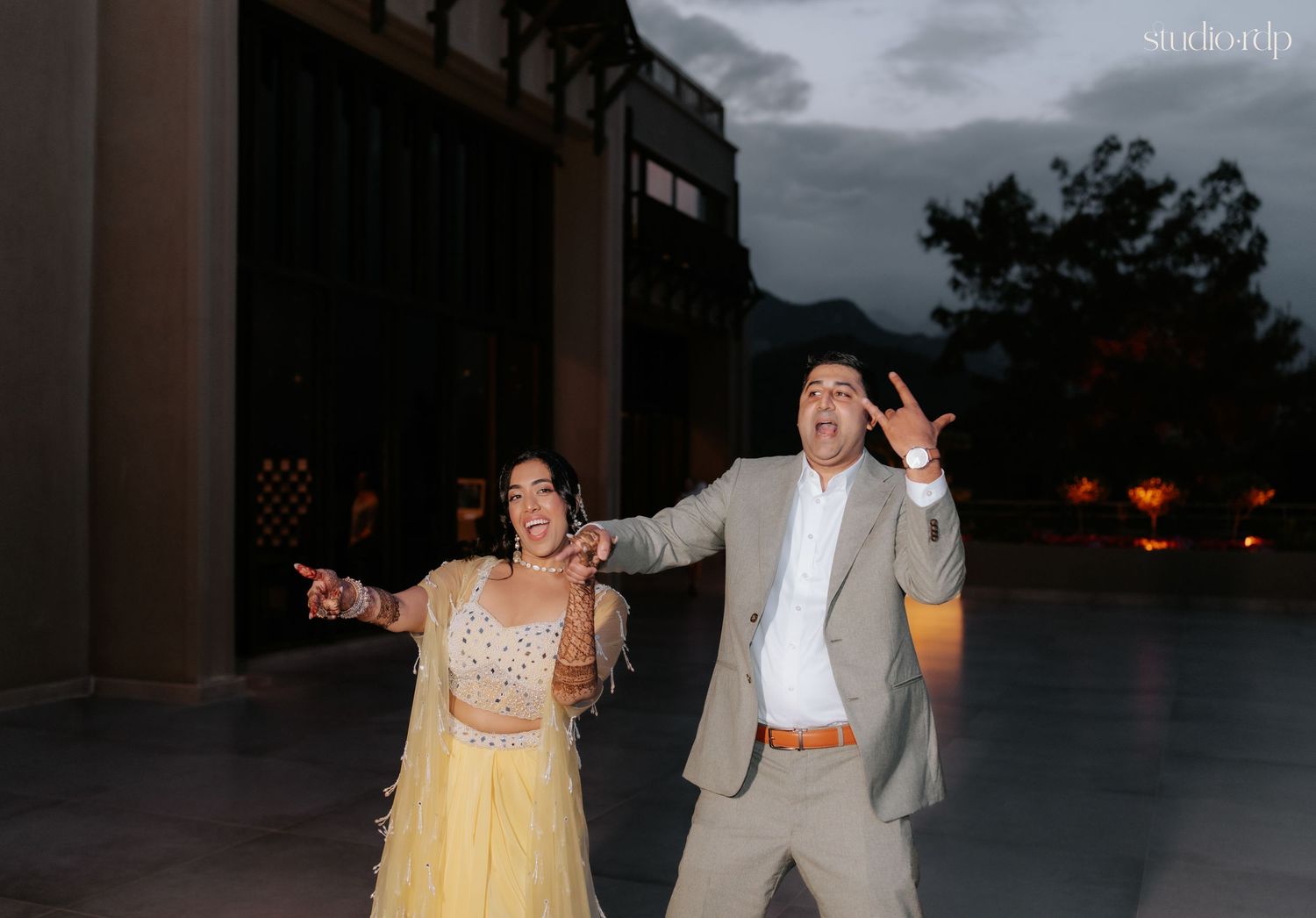 Couple dancing joyfully outdoors at Indian wedding reception, woman in yellow lehenga, man in gray suit, mountains backdrop.