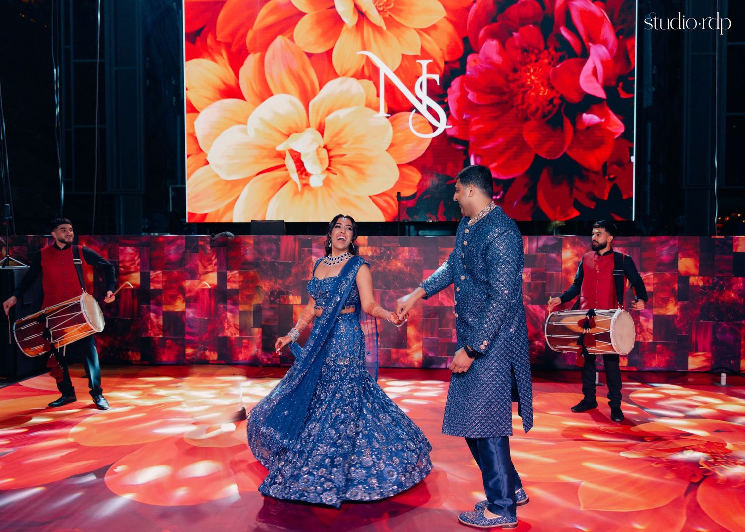 Couple dancing in blue embroidered outfits at a vibrant Indian wedding sangeet with drummers and floral LED backdrop.