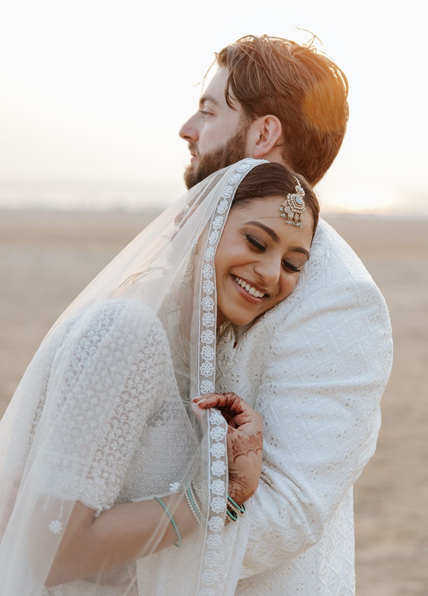 A bride in white embroidered lehenga with maang tikka embraces groom at golden hour in a romantic wedding portrait.