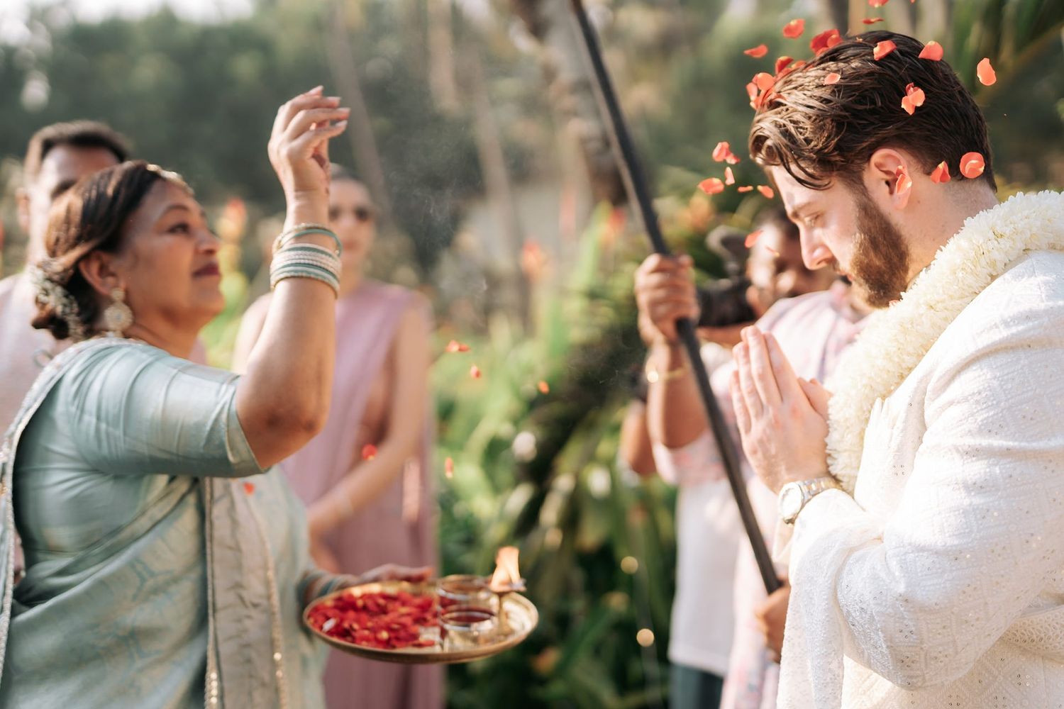 Indian wedding ceremony with groom in white sherwani receiving blessings from elder woman throwing flower petals.