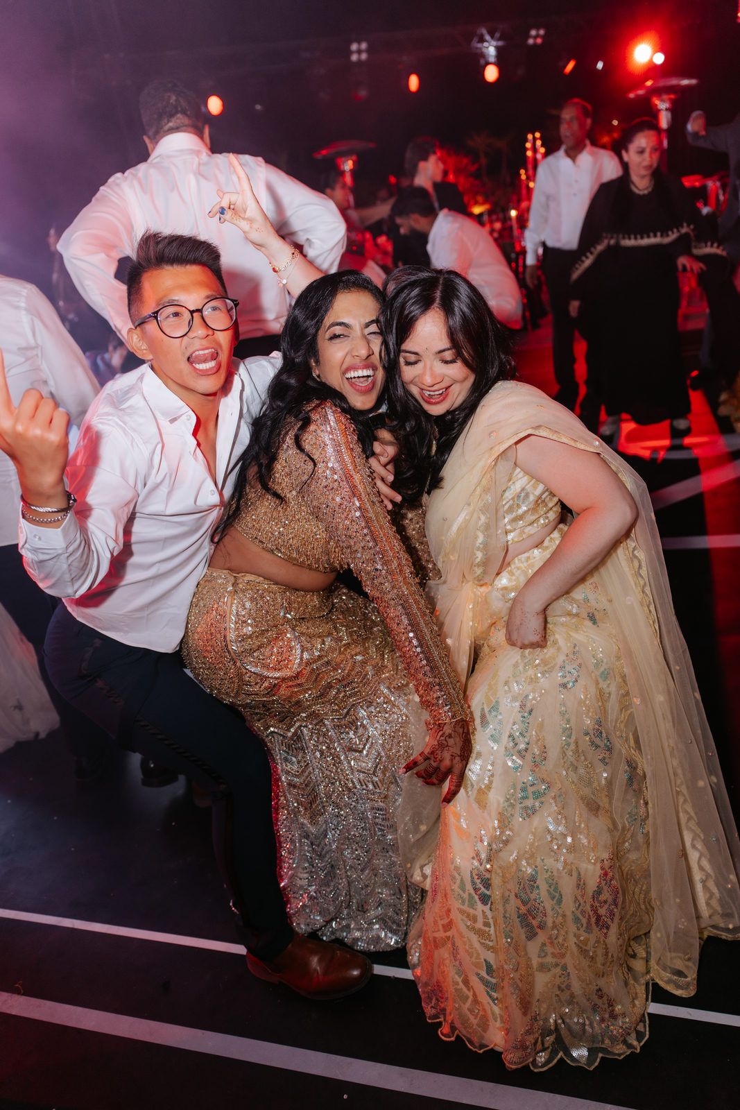 Three friends pose and dance together in sparkly traditional outfits at a lively wedding reception party.