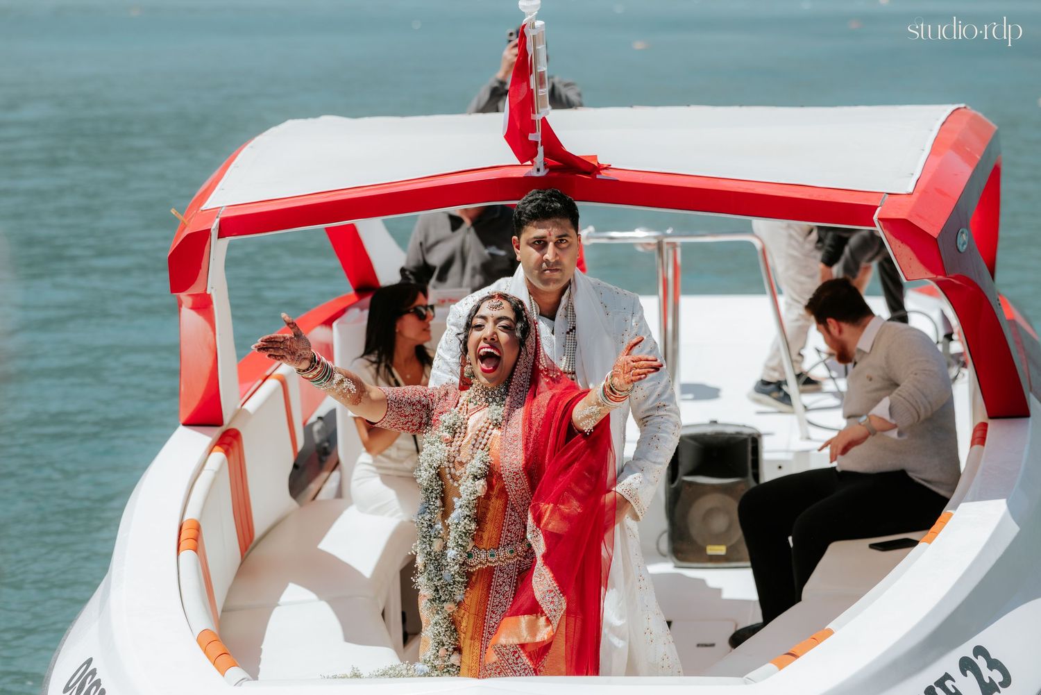 Bride and groom celebrating on a white speedboat on turquoise water, wearing vibrant Indian wedding attire.