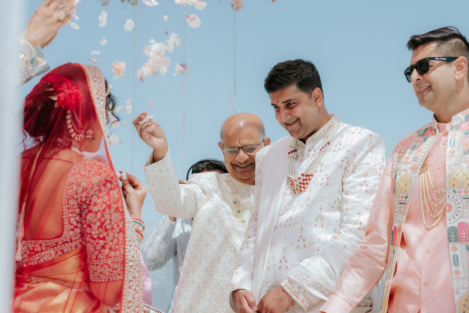 Indian groom in white sherwani exchanges flower garlands with bride during jaimala ritual at outdoor wedding.