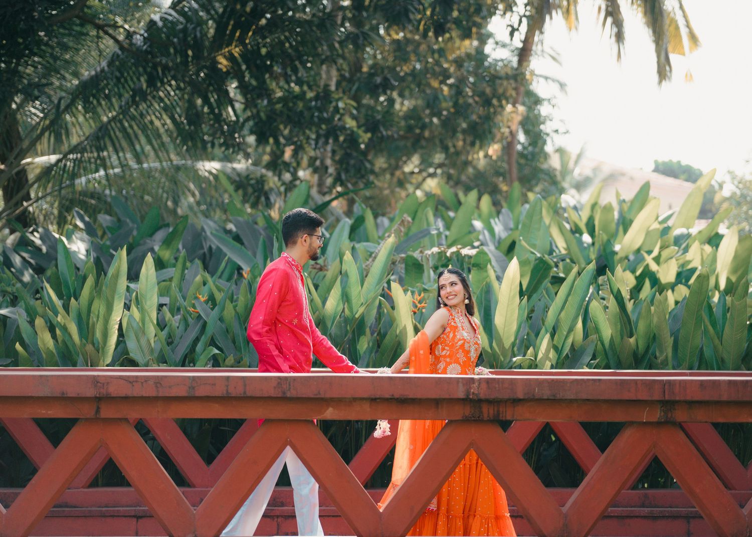 Two people in vibrant red and orange traditional attire sit together on a wooden bridge surrounded by tropical foliage.
