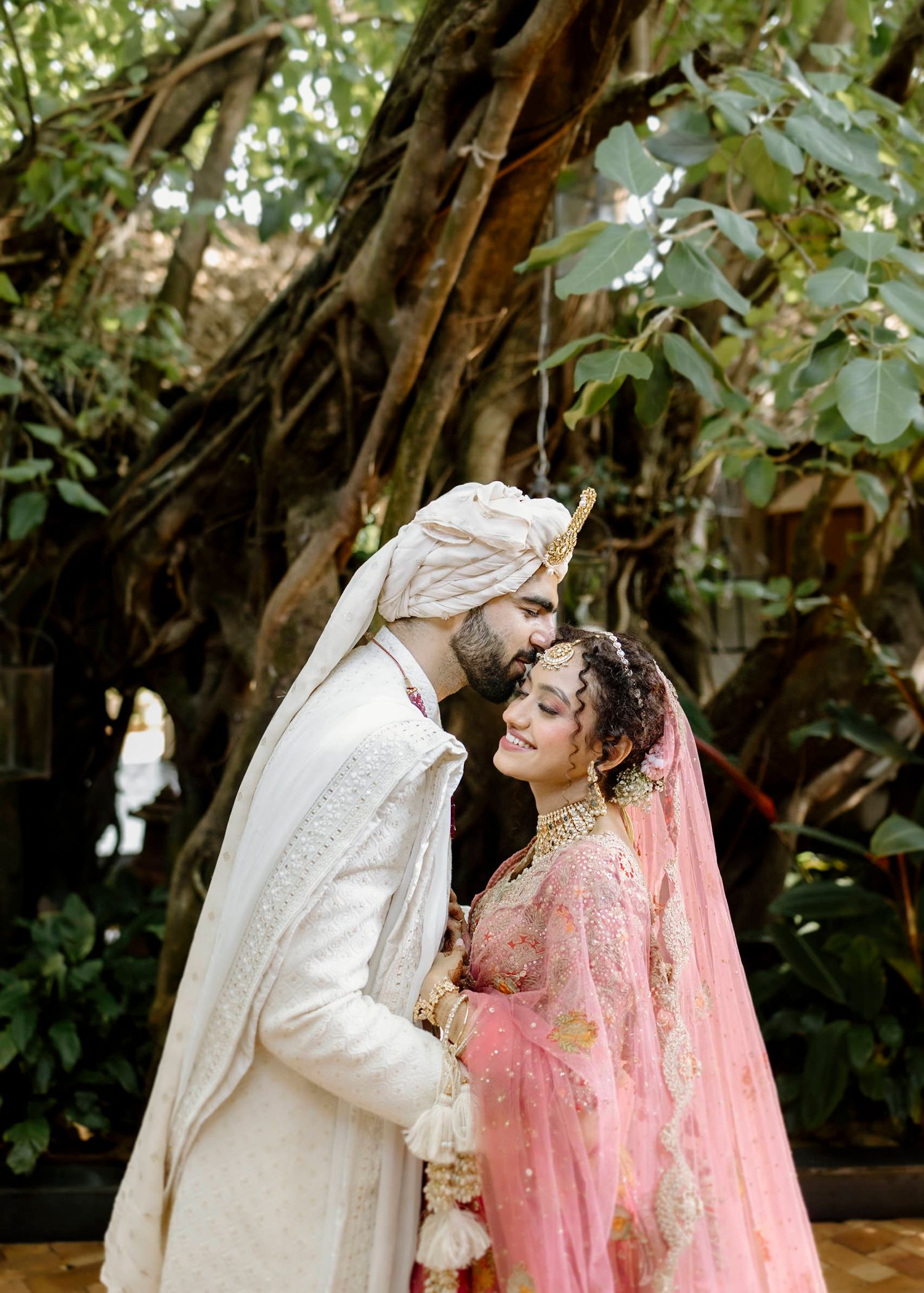 A couple in traditional South Asian wedding attire share an intimate moment in a lush garden setting.