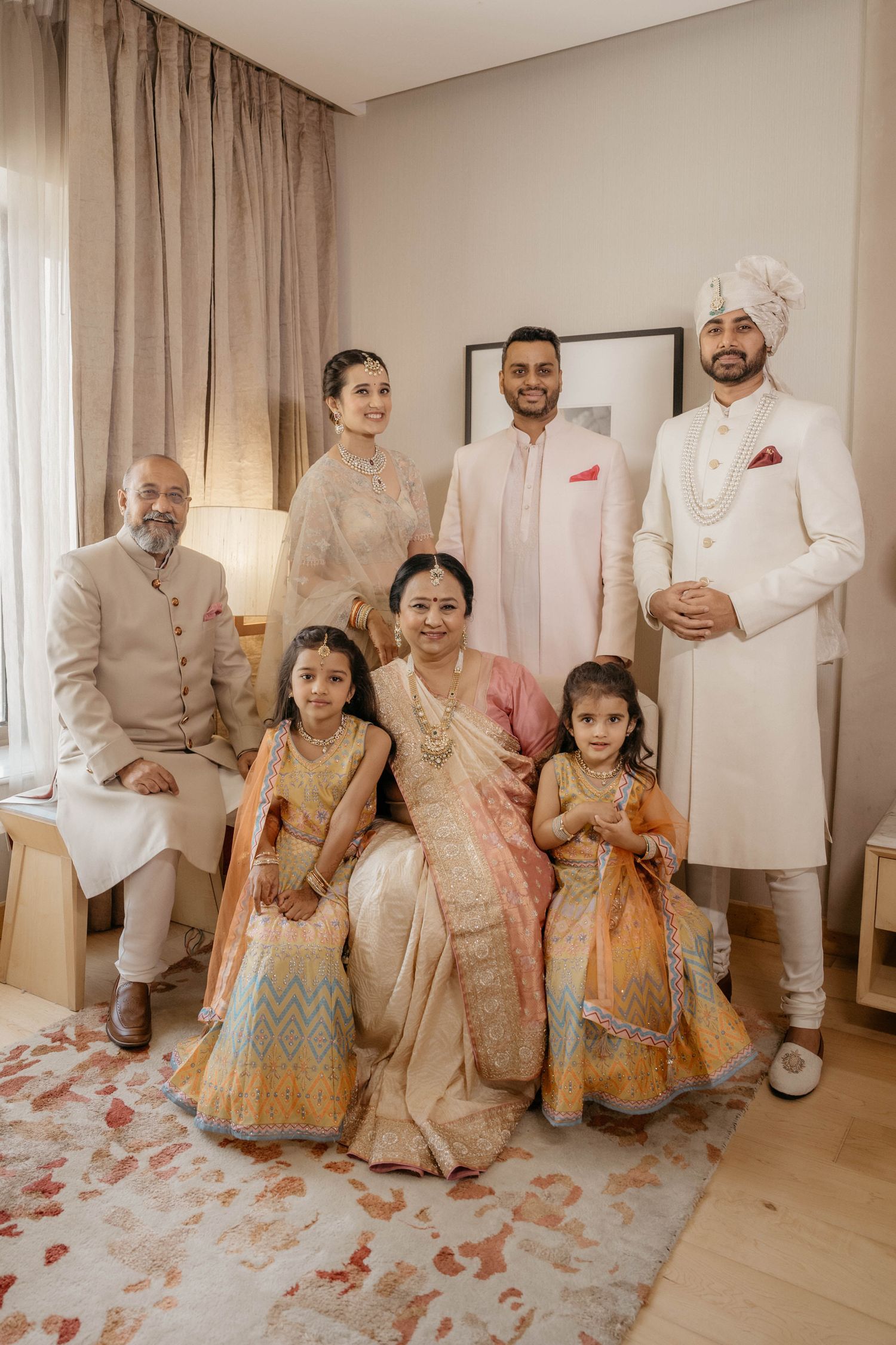 A family dressed in elegant Indian traditional attire poses together in a living room with beige curtains and decor.