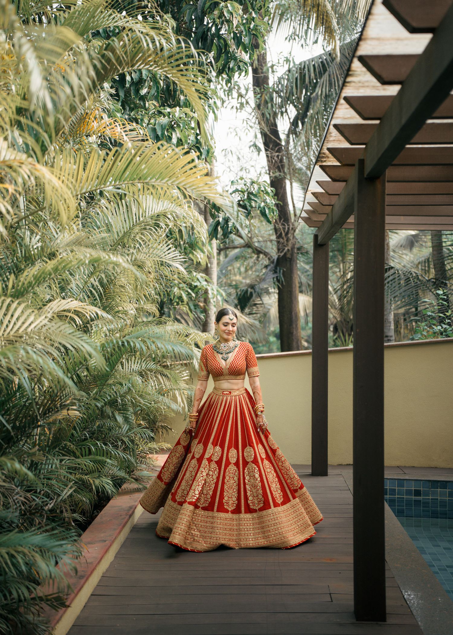 Beautiful red and gold embroidered lehenga captured against lush tropical palm trees and wooden pergola by a pool.