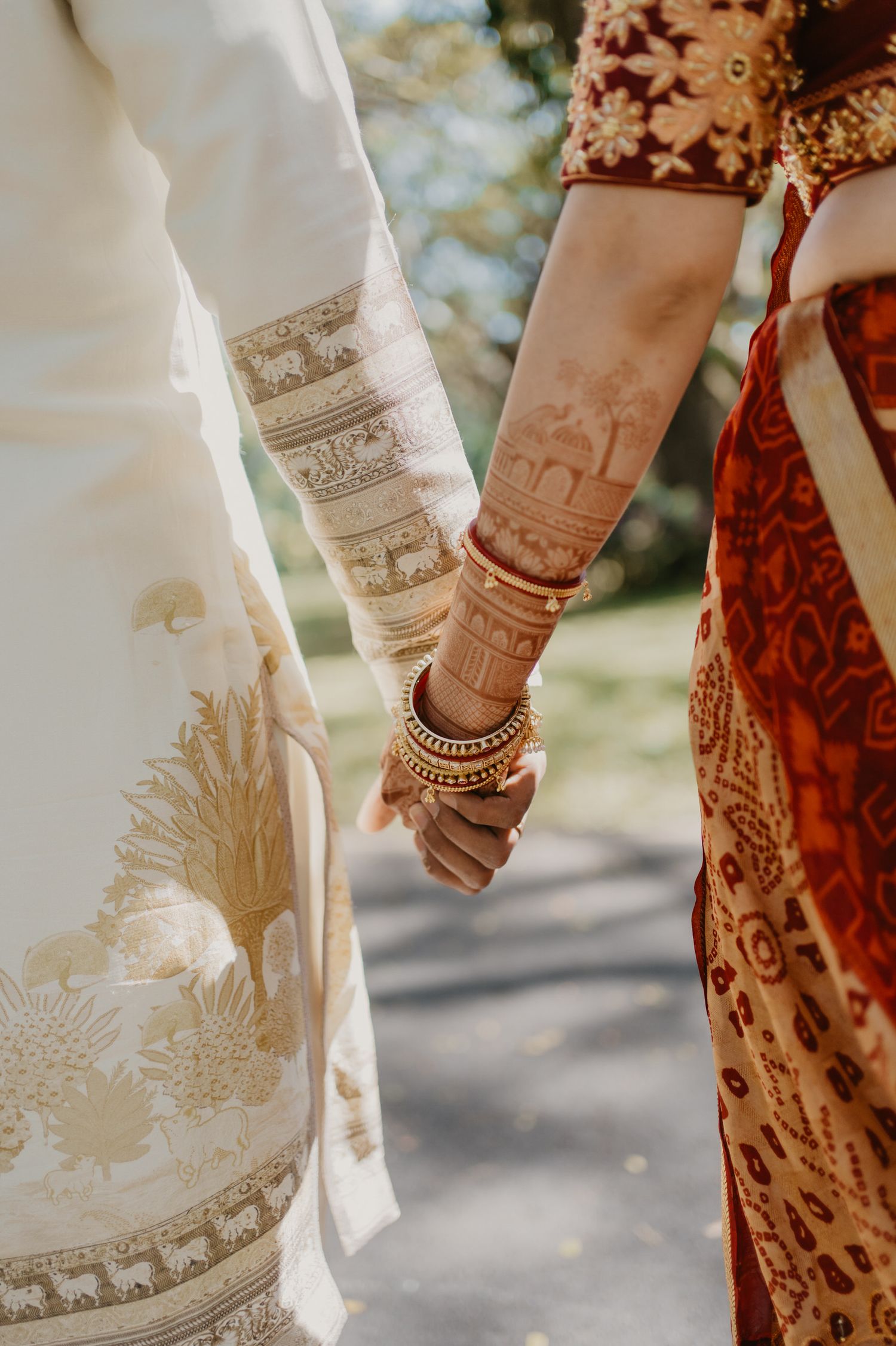 Close-up of hands with ornate henna designs and bangles as a couple holds hands in traditional Indian wedding attire.