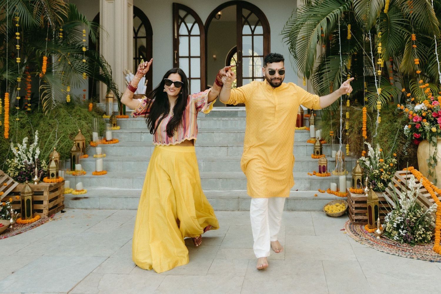 Brightly dressed people walk together in a spacious indoor venue adorned with potted plants and arched windows.