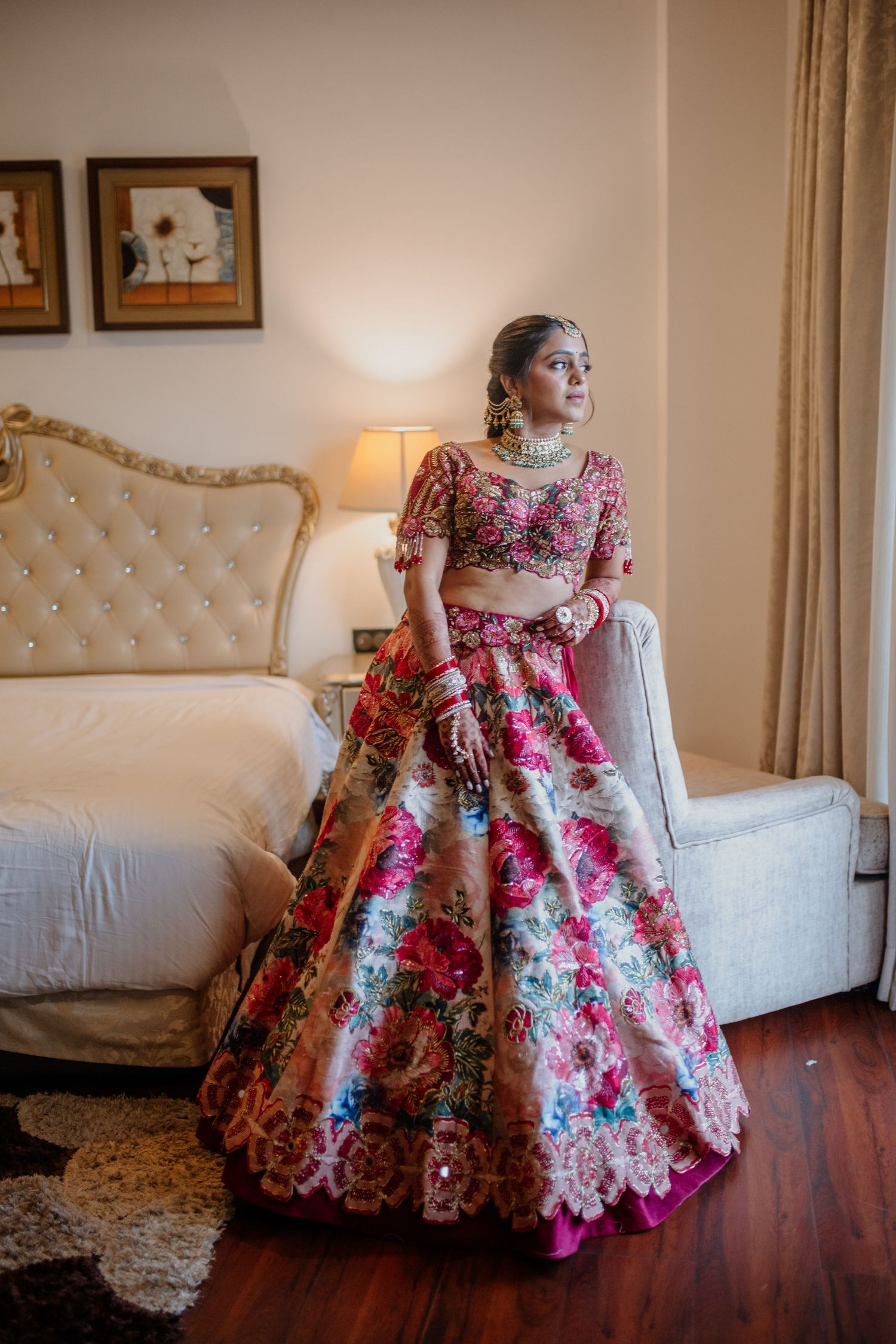 A person in a stunning floral pink ball gown with lace details poses gracefully on a hardwood floor by a cream sofa.