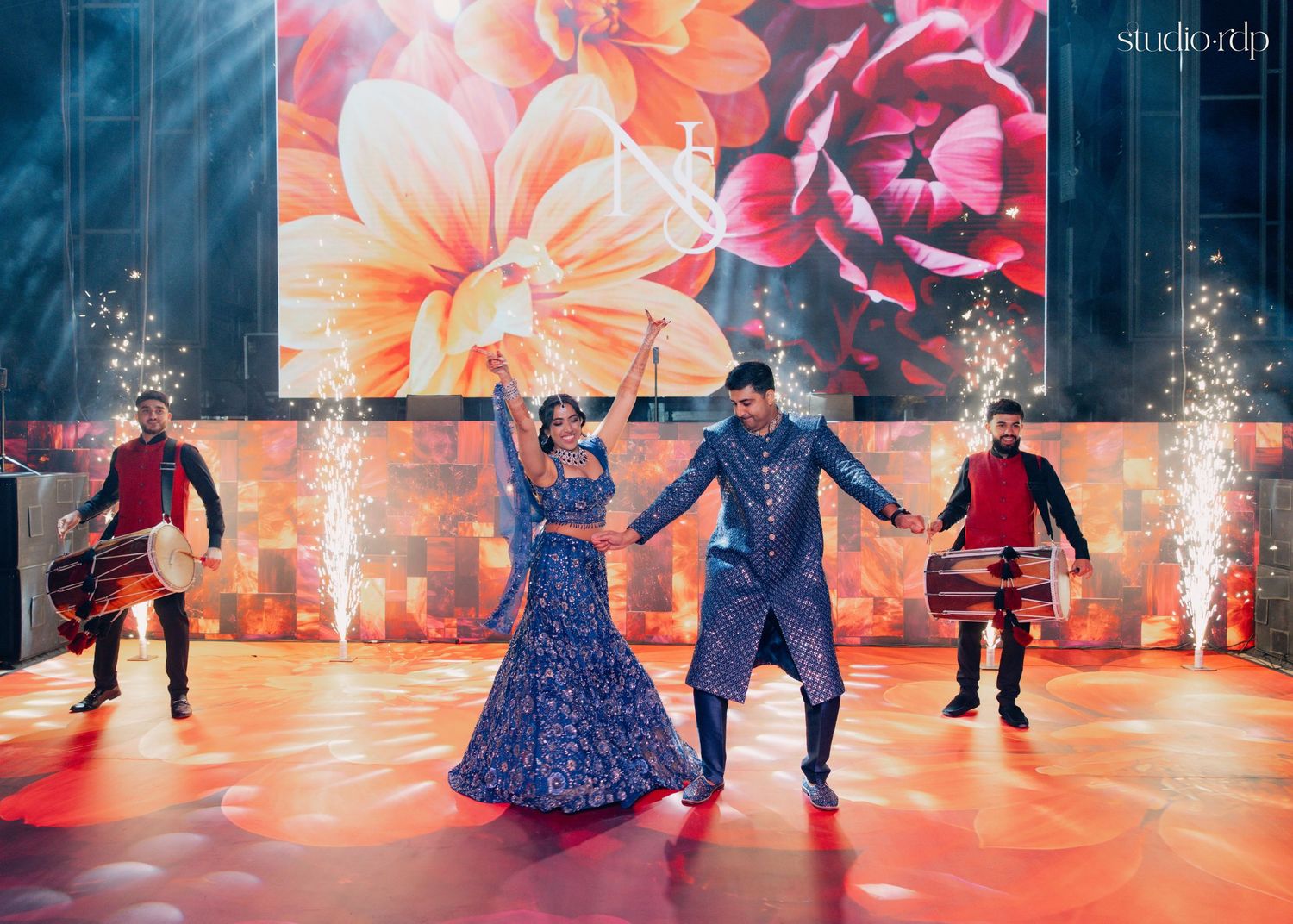 Couple dancing on stage in blue outfits at Indian wedding with fireworks and floral backdrop.
