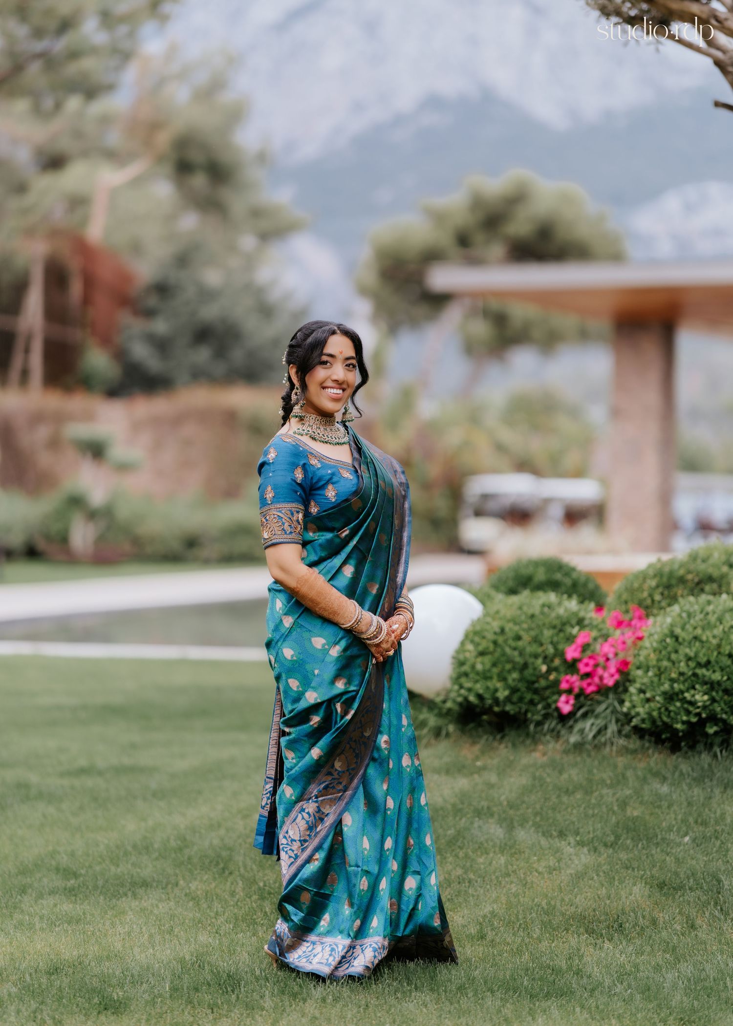 Woman in teal embroidered saree with gold jewelry smiling outdoors with mountains in background.