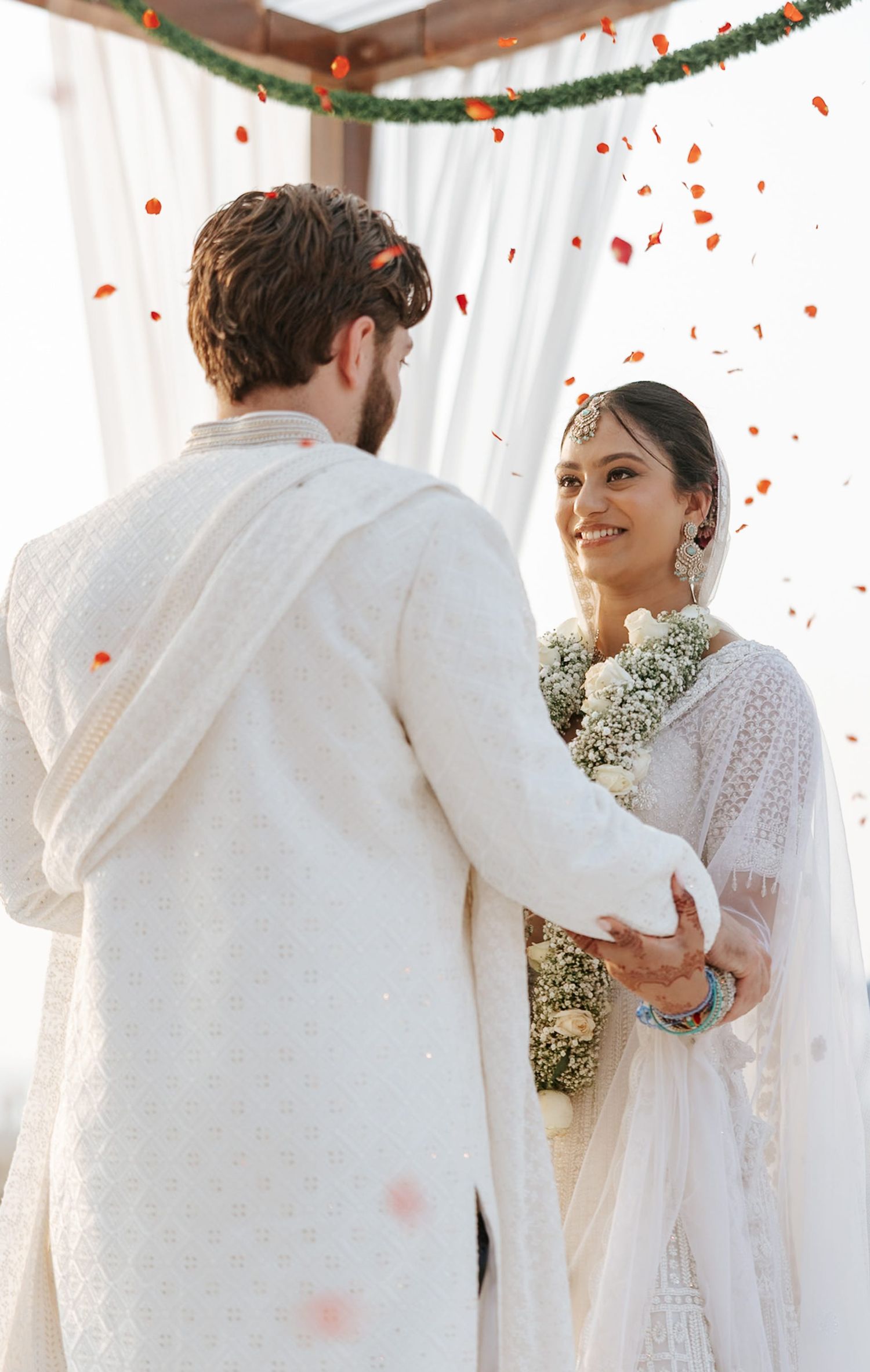 Couple in white traditional attire exchange vows at an outdoor wedding ceremony with flower petals falling.