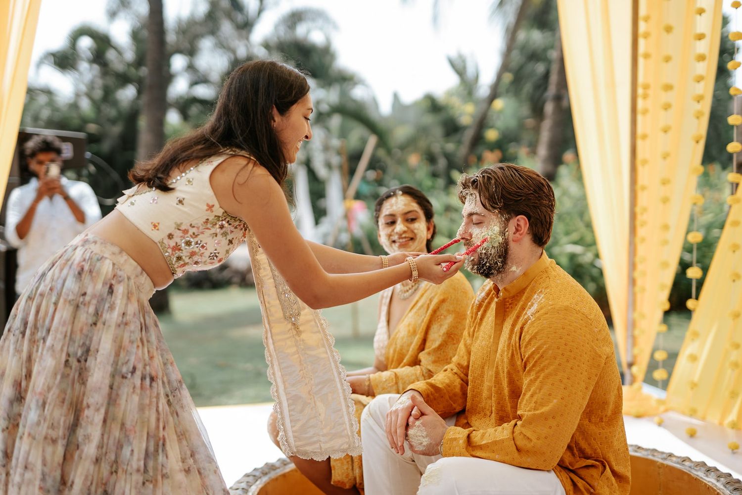 A woman in floral lehenga applies haldi to a bearded groom in yellow kurta at an outdoor Indian wedding ceremony.