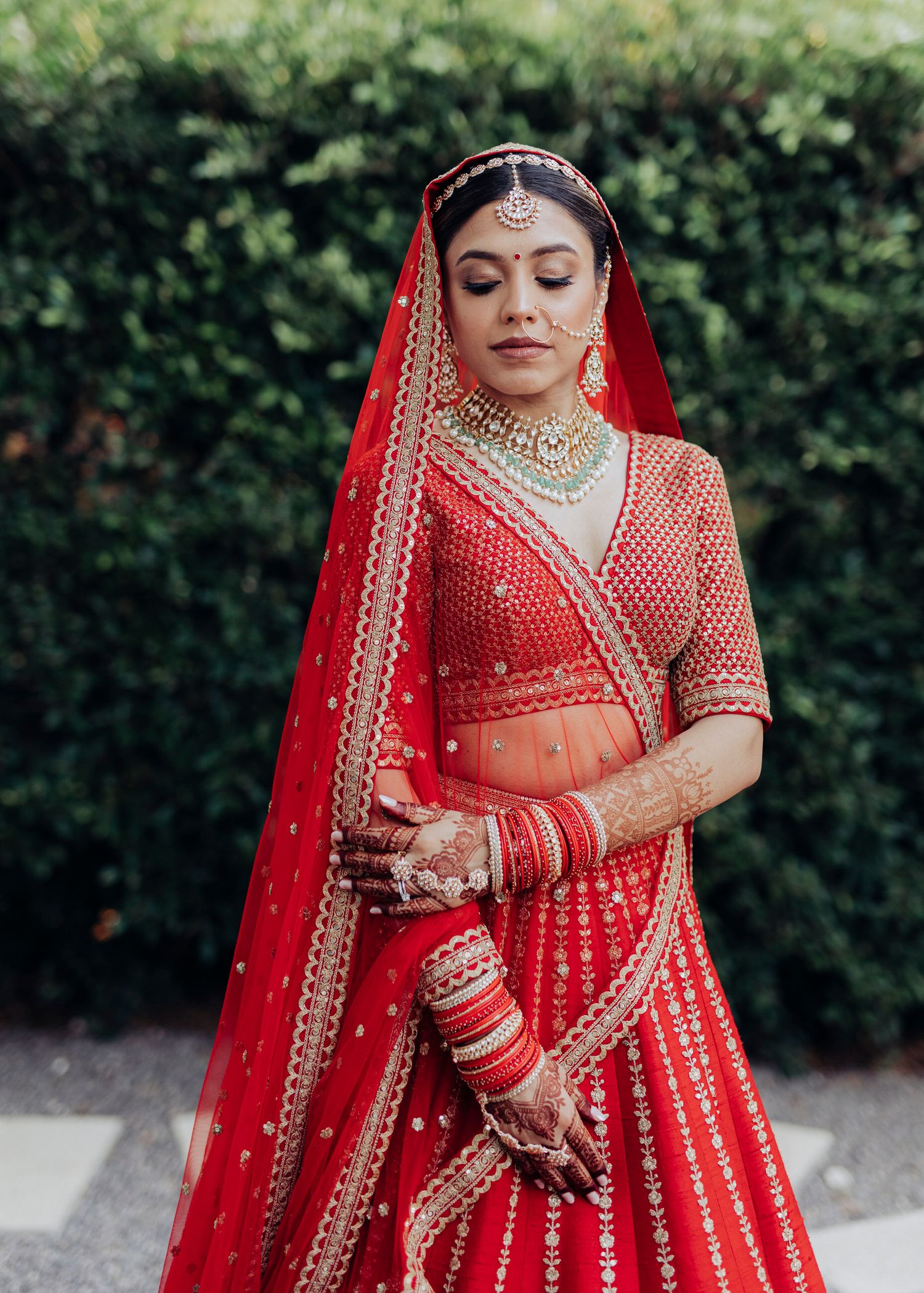 A traditional Indian bride in an ornate red and gold embroidered lehenga ensemble adjusts her dupatta and jewelry.