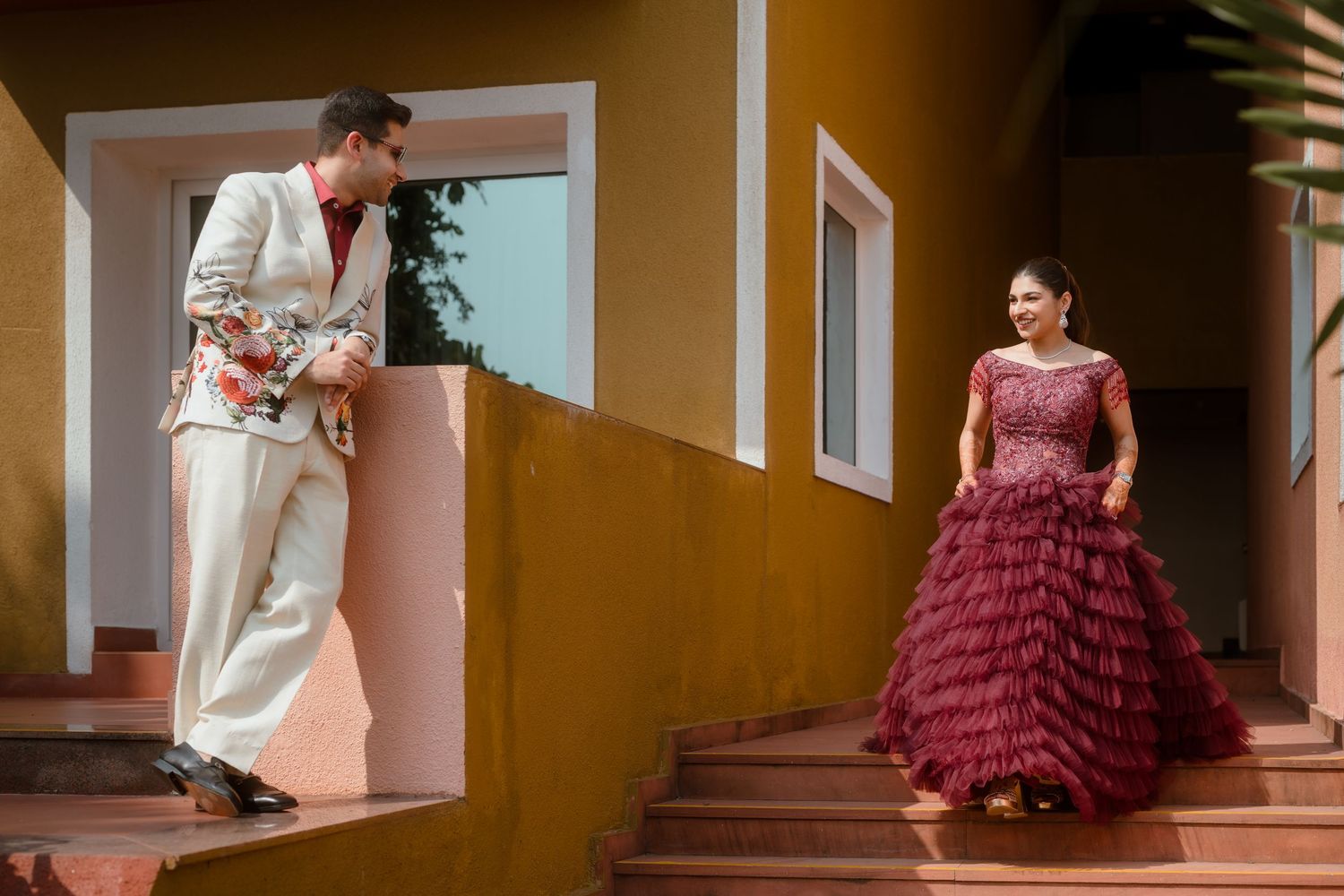 A model in a burgundy ruffled gown poses elegantly on a staircase with warm-toned walls.