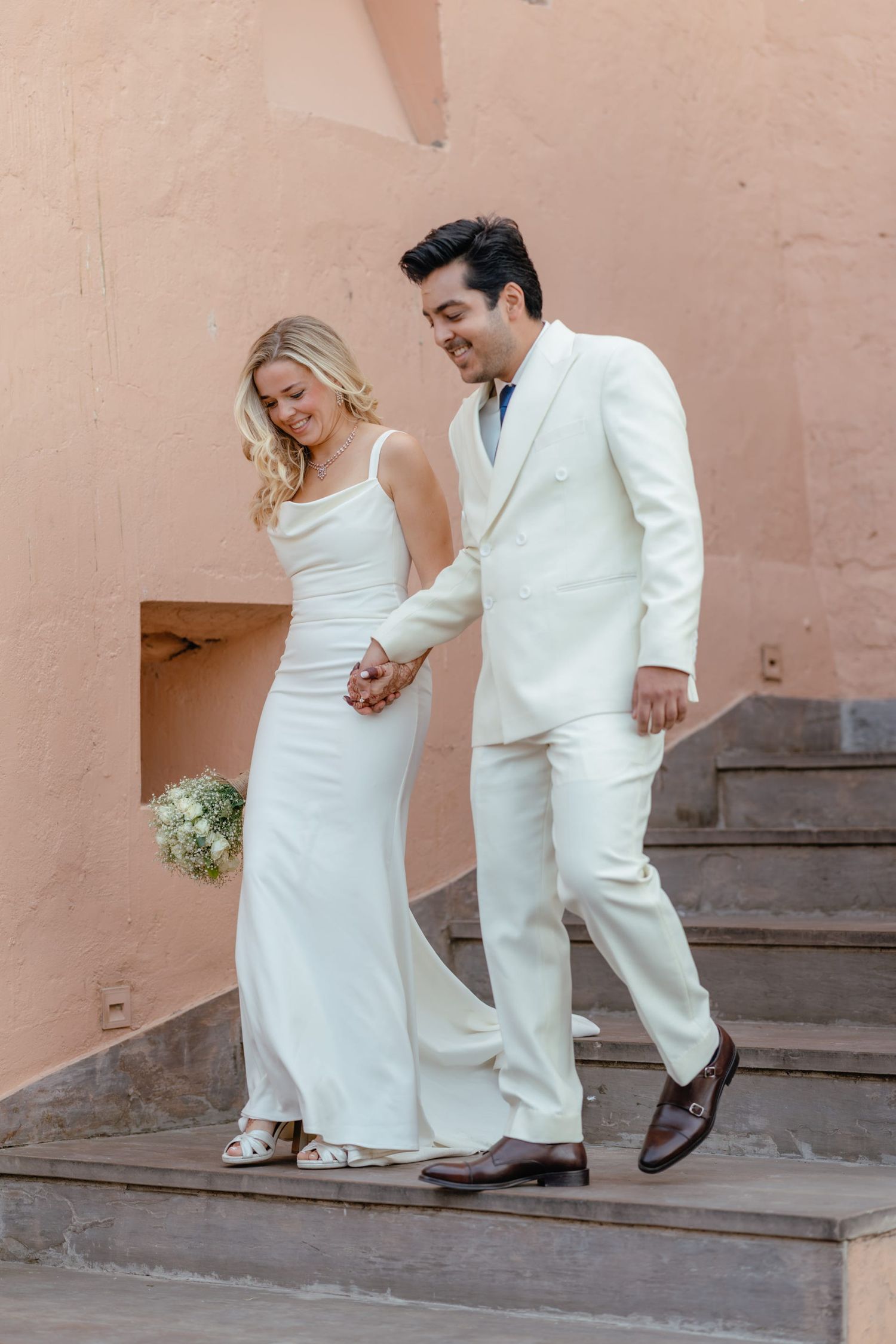A couple in white wedding attire walking up stone steps together in an elegant setting.