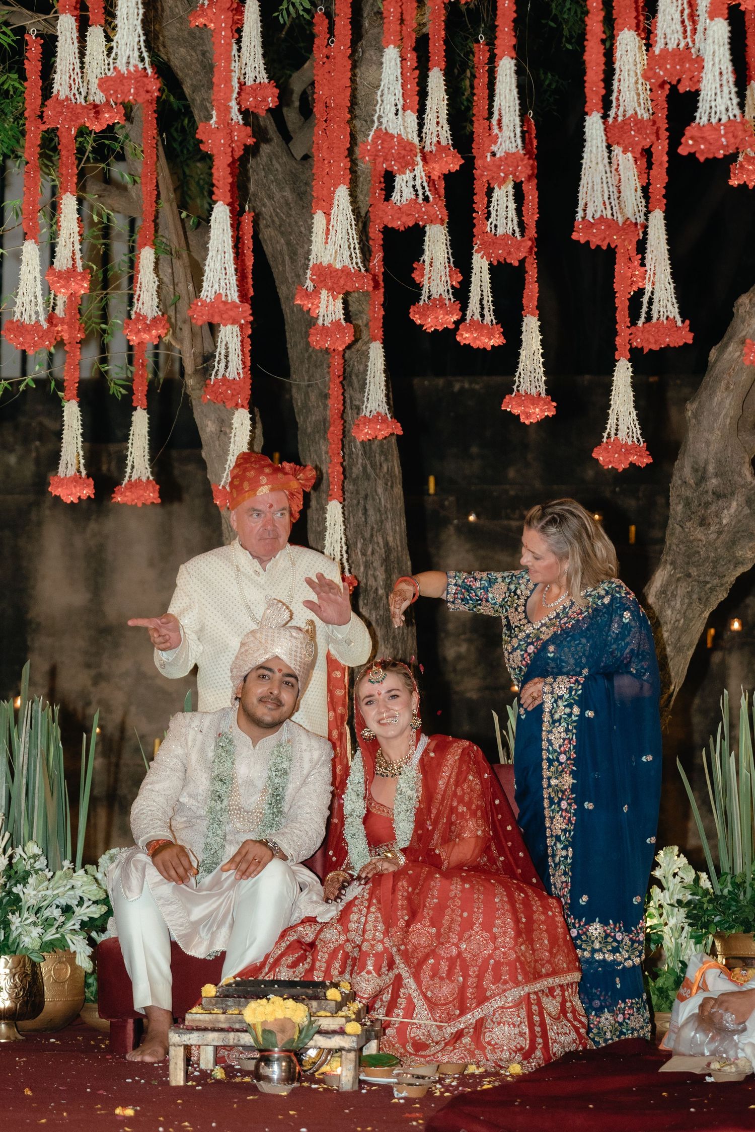 Family gathering during wedding ceremony under decorative hanging flowers with traditional rituals.