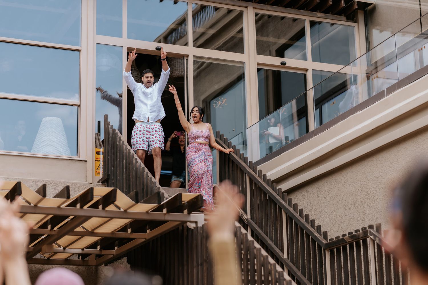 Couple celebrating on outdoor staircase at wedding venue, guests cheering below.